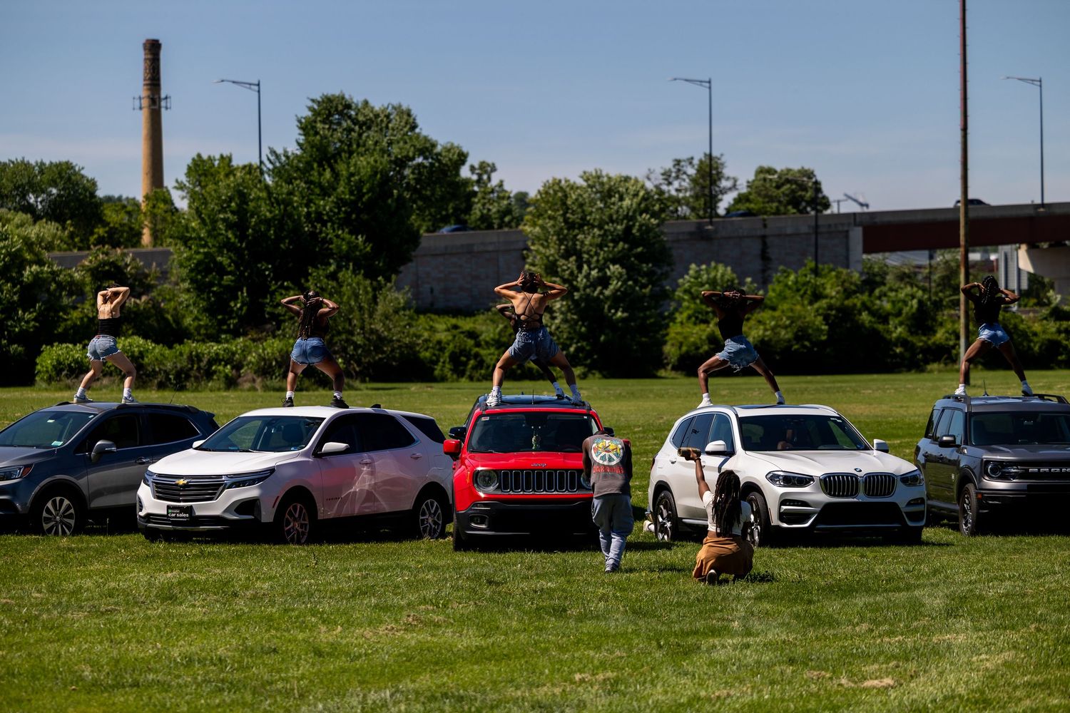 Cheerleaders perform acrobatic stunts outdoors near parked SUVs and cars on a green field with trees in background.
