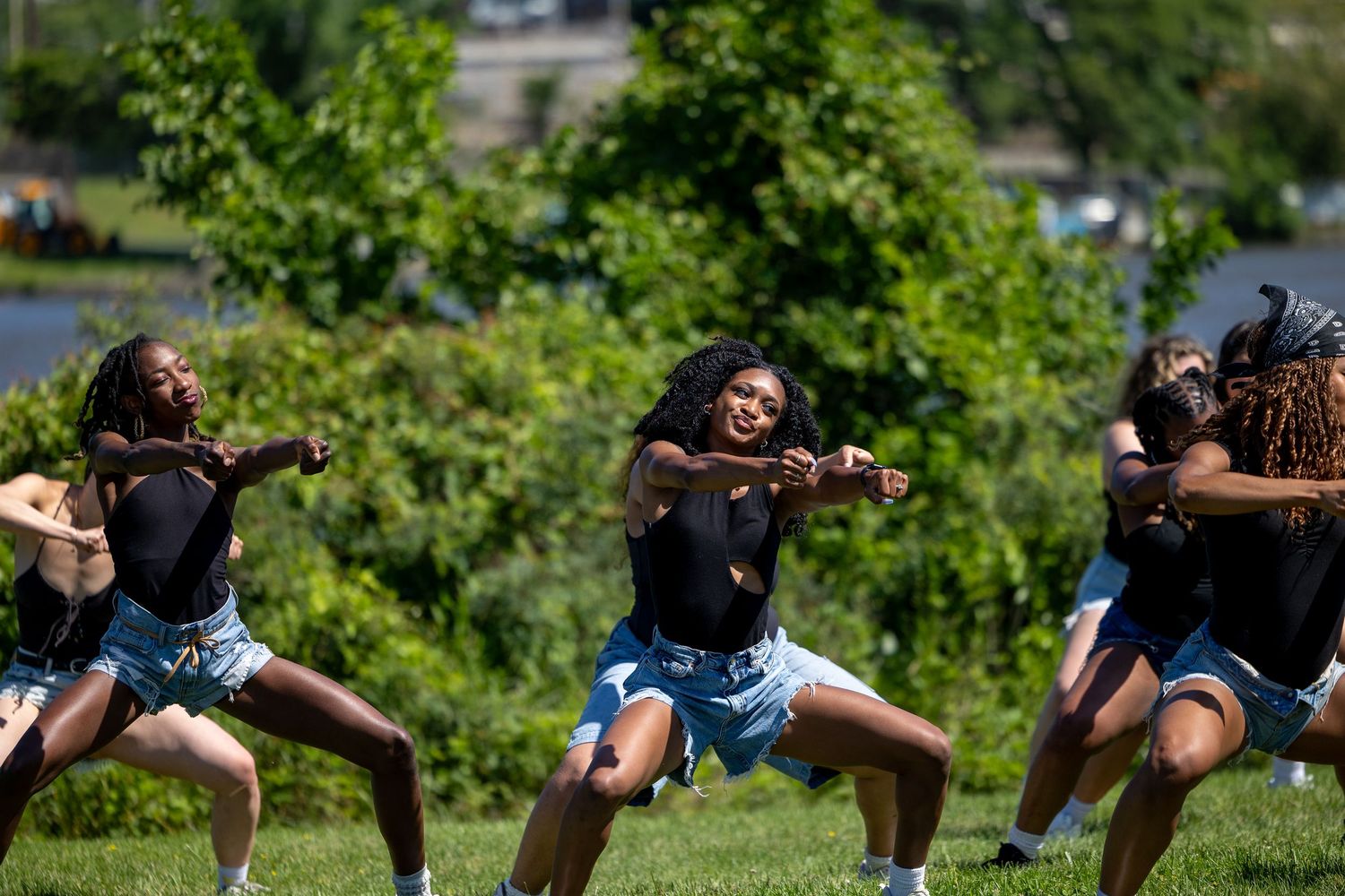 Young women performing an energetic dance routine outdoors on green grass in matching black tops and denim shorts.