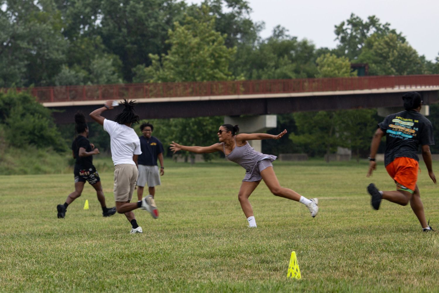 Group of young people playing flag football on a green field outdoors near a bridge on a cloudy day.