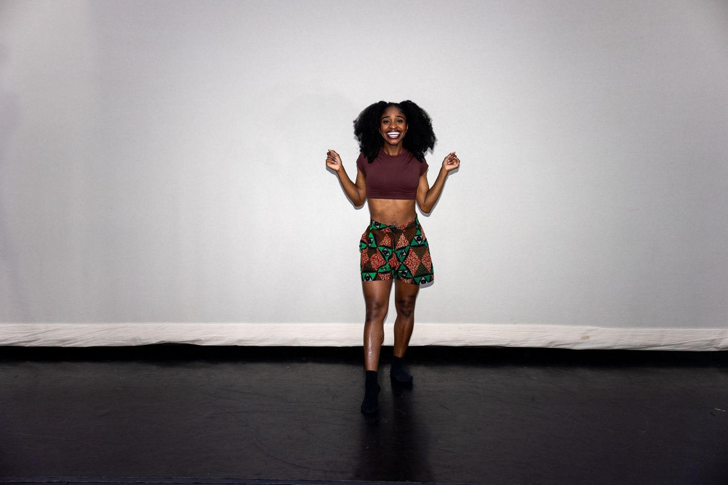 Energetic Black woman in African print skirt and maroon top dancing joyfully on a dark stage.