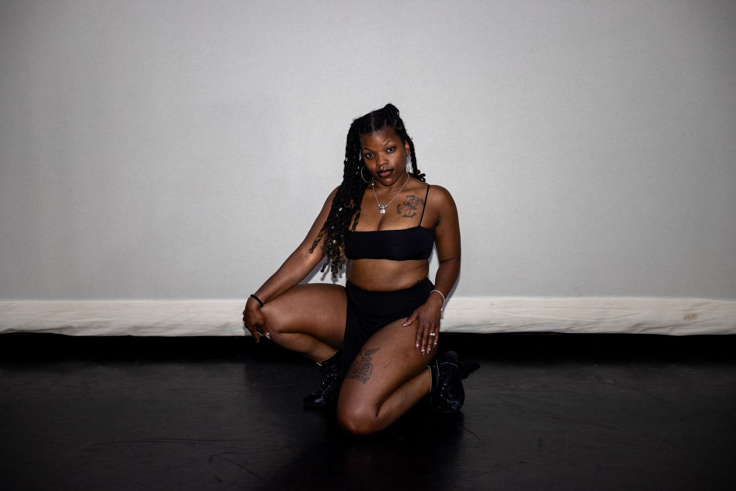 Black woman in black bikini set poses gracefully on studio floor with long braids.