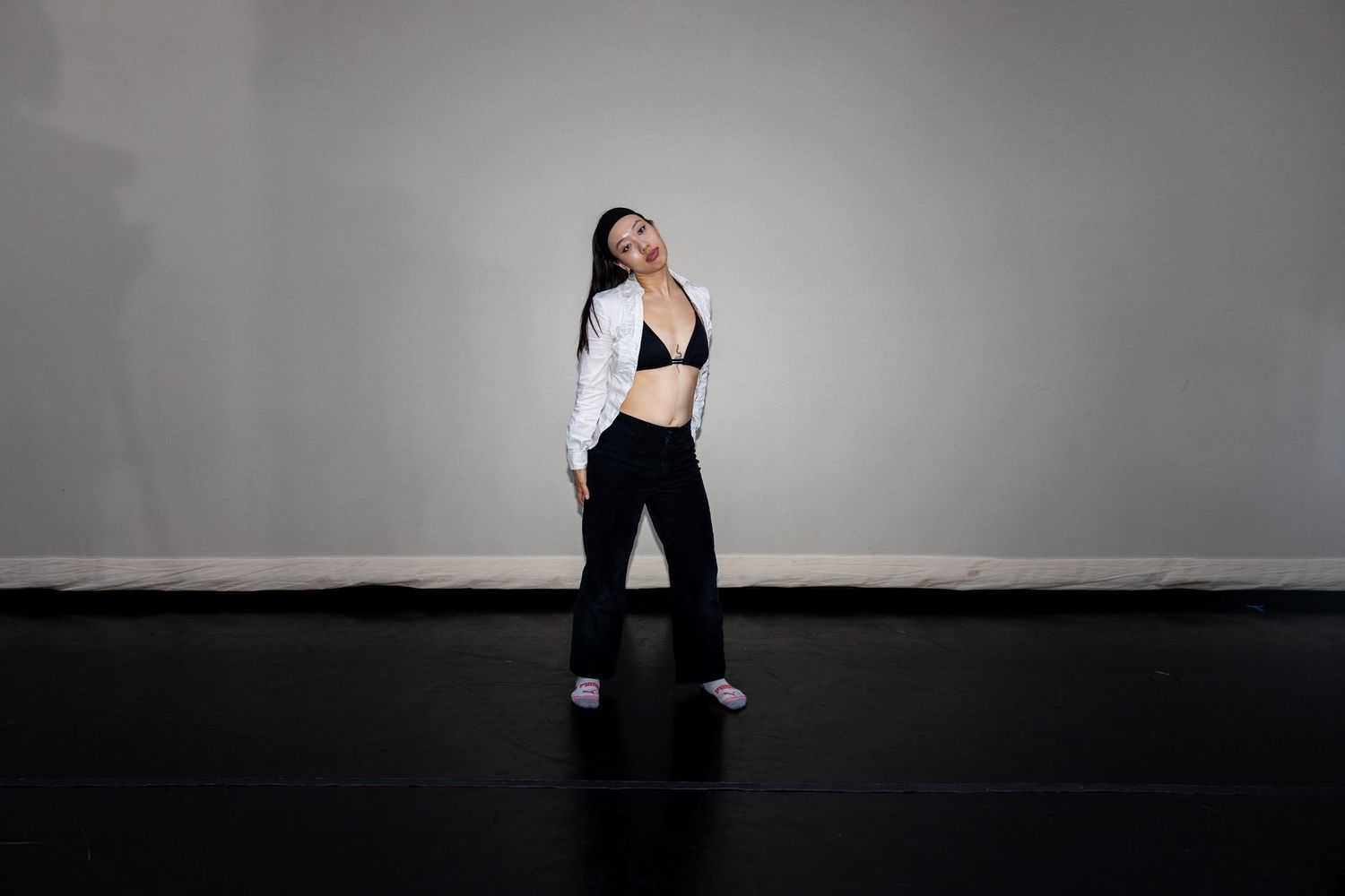 A woman poses confidently in a black sports bra, white jacket, and black pants on a dark studio floor.