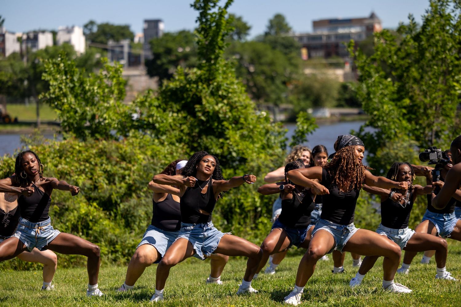 Group of dancers performing synchronized routine outdoors on green lawn near river on sunny day.