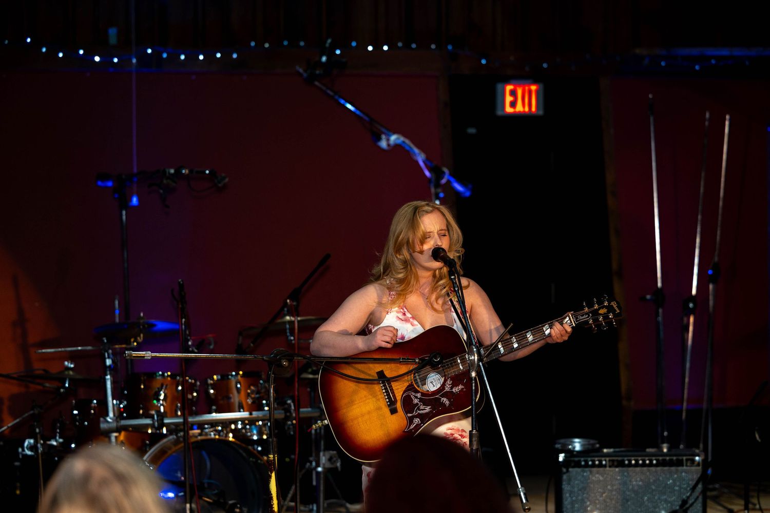 Female guitarist performing on stage with acoustic guitar, drum kit visible behind her, under warm stage lighting.