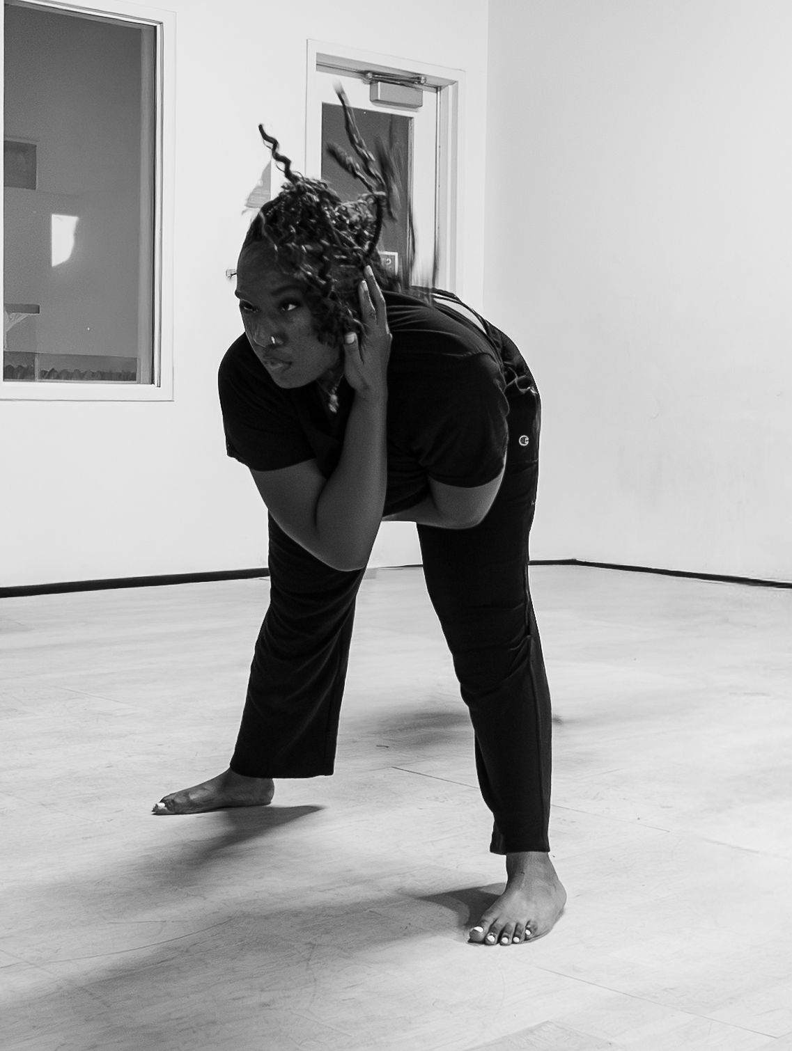 Black and white photo of a dancer bending forward with decorative hair accessories in a white studio.