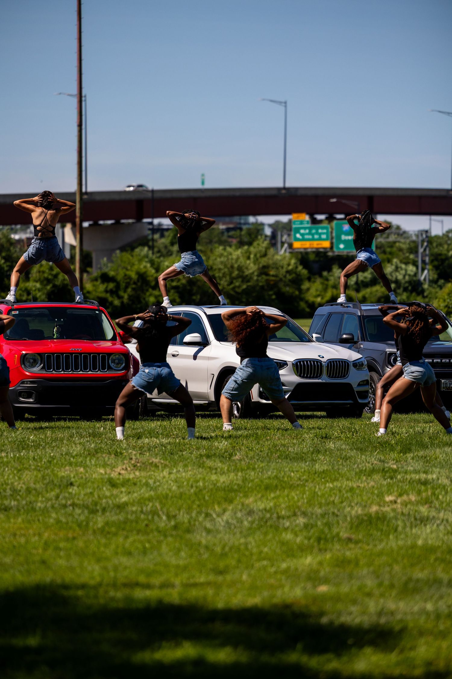 Dancers performing acrobatic moves on grass near parked cars including a red Jeep and white BMW on a sunny day.