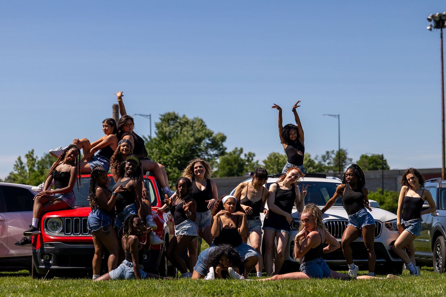 Group of young women posing outdoors near cars on a sunny day, smiling and raising arms in a joyful celebration.