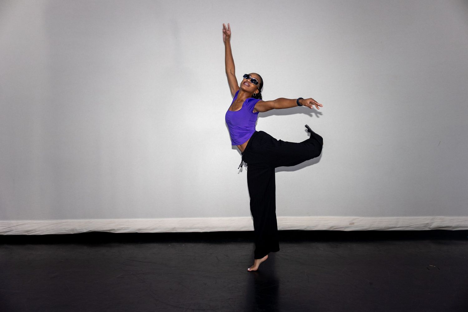 A dancer in a purple top and black pants performs an elegant arabesque in a studio.