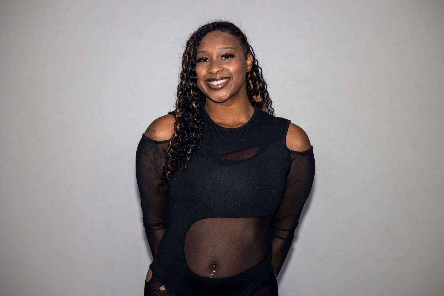 A plus-size woman with braided hair smiles in a black mesh cutout top against a dark background.