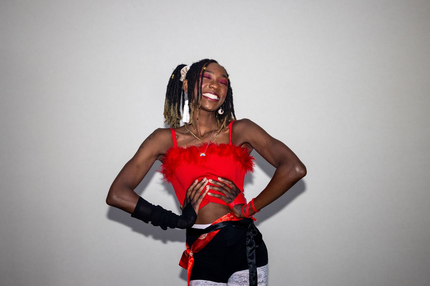 Joyful performer in red feathered crop top and black gloves laughing with hands on hips against gray background.