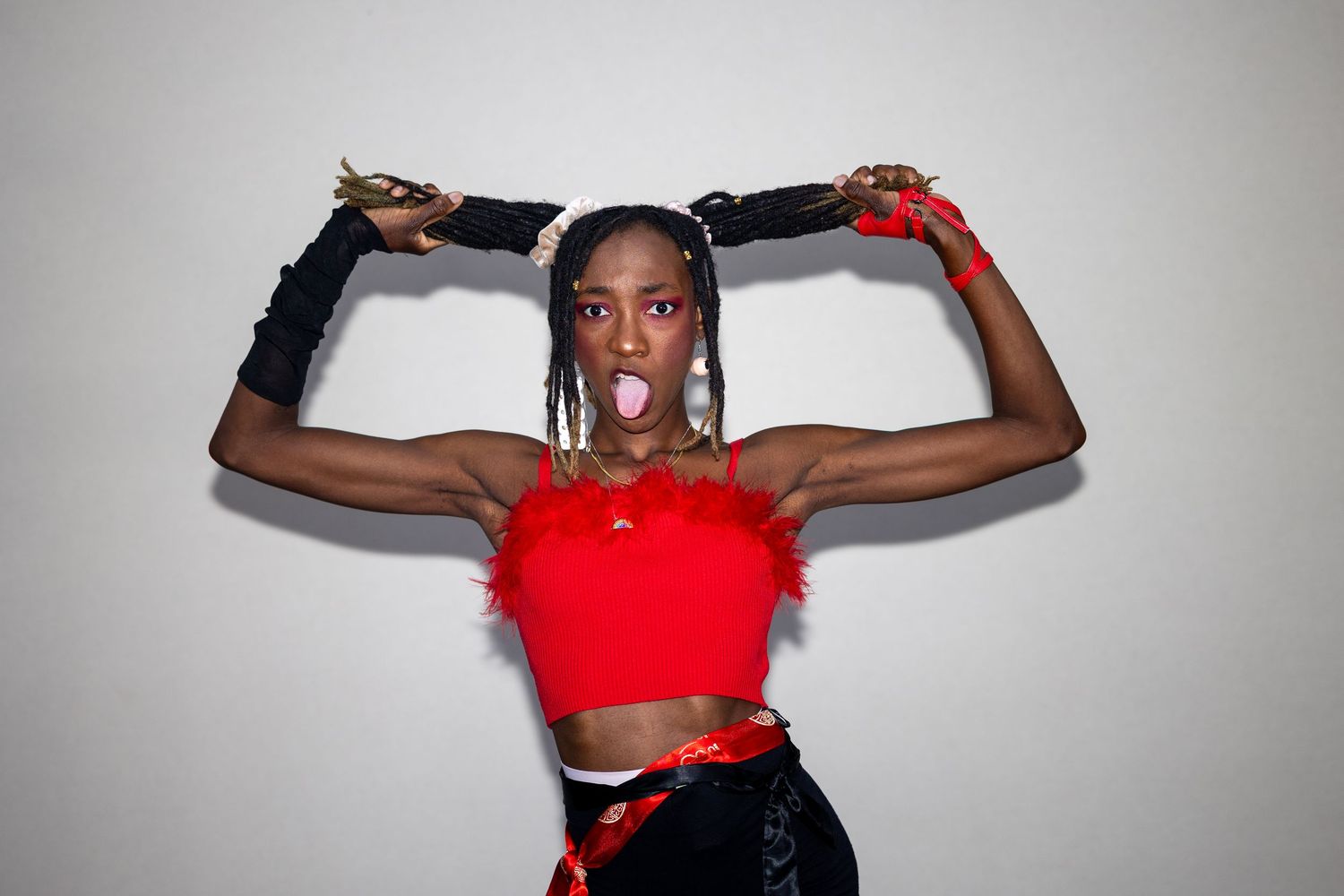 Young Black woman in red crop top and black gloves striking a fierce pose with pigtails raised overhead.