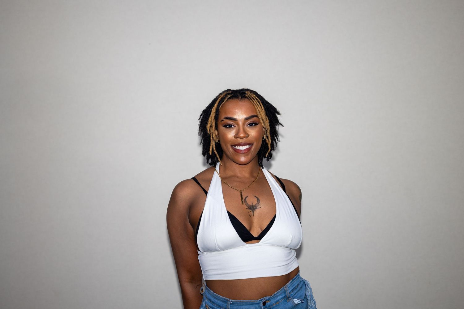 Confident young woman with braids in a white halter crop top and denim jeans smiling brightly.