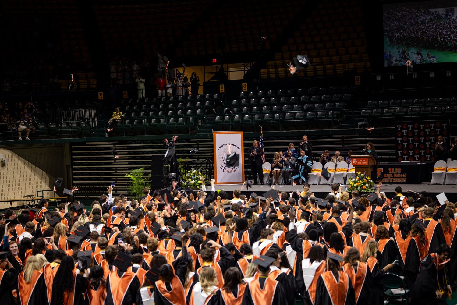 Large graduation ceremony crowd in orange and black regalia fills an indoor arena.