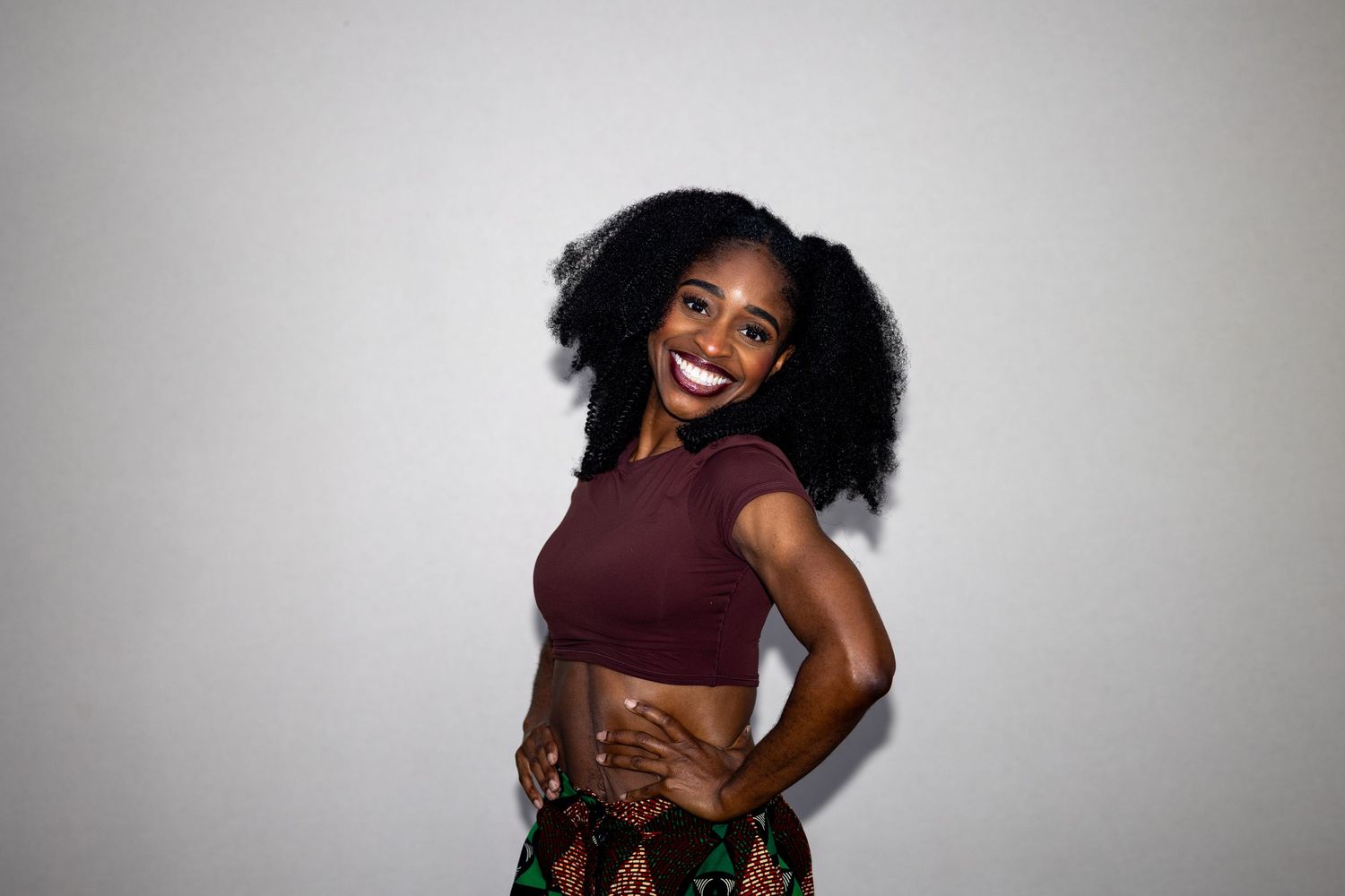 Smiling woman in maroon crop top and patterned skirt poses with hands on hips against white background.
