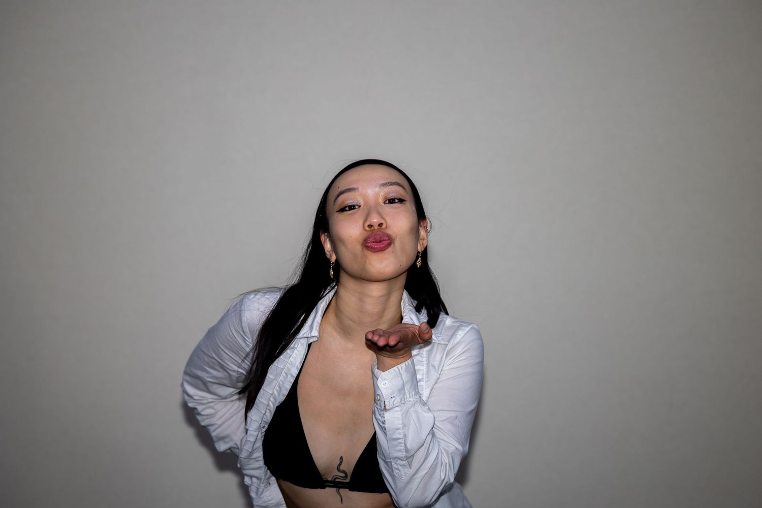 Young Asian woman in white jacket and black bralette blows a kiss toward the camera.