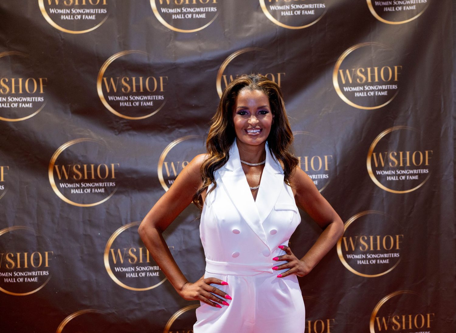 Woman in white sleeveless blazer dress poses confidently on WSHOF red carpet event backdrop.