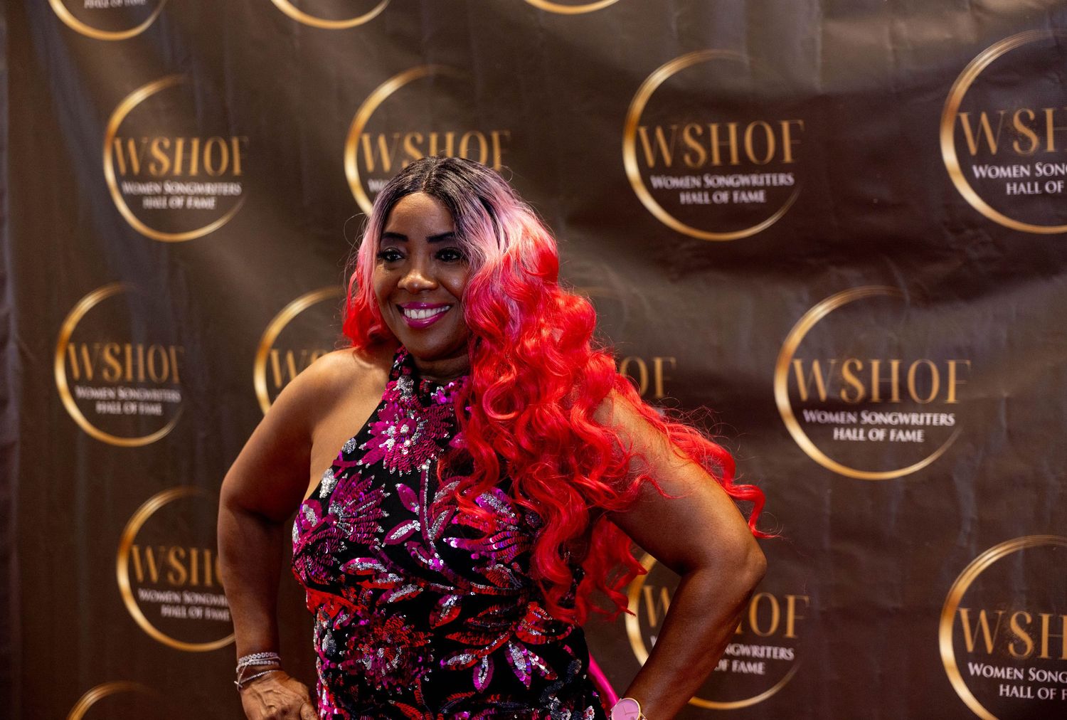 Woman in vibrant red wig and floral dress poses on WSHOF Women Songwriters Hall of Fame red carpet event.