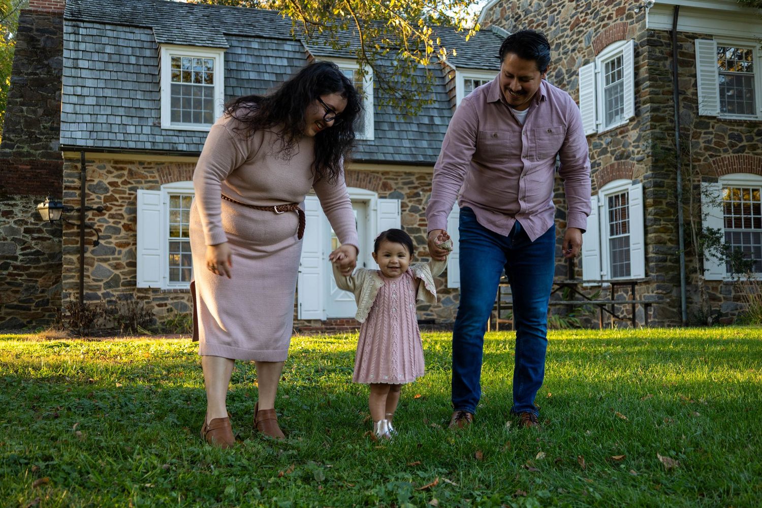 Family of three walking on green lawn in front of stone house, parents holding toddler girl's hands outdoors.