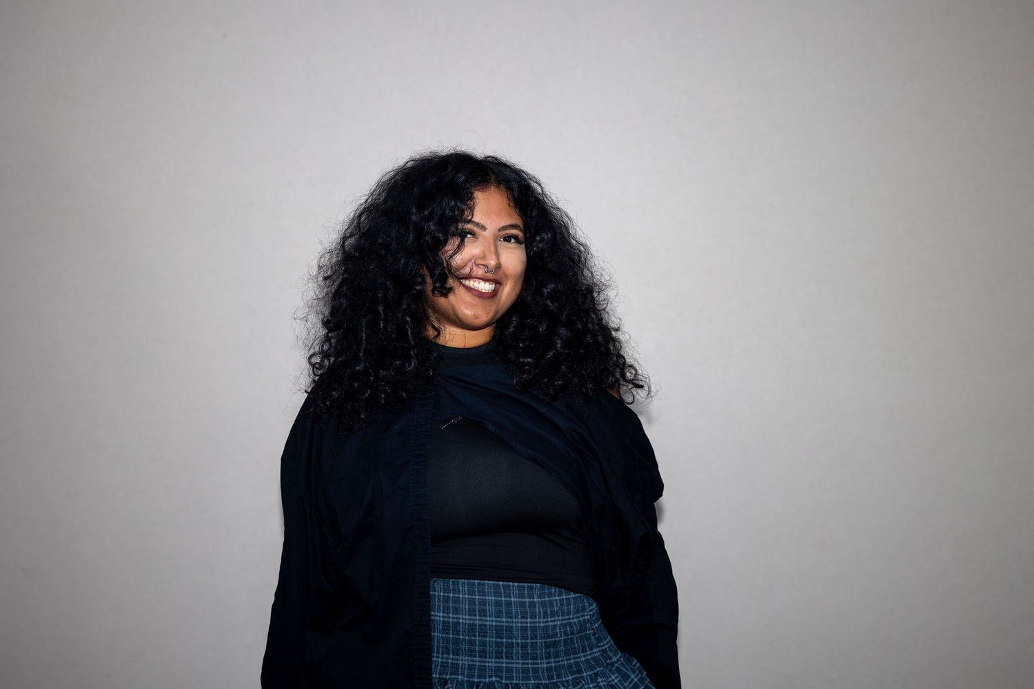 Smiling woman with long curly hair wearing a black top and plaid skirt posing against a gray background.