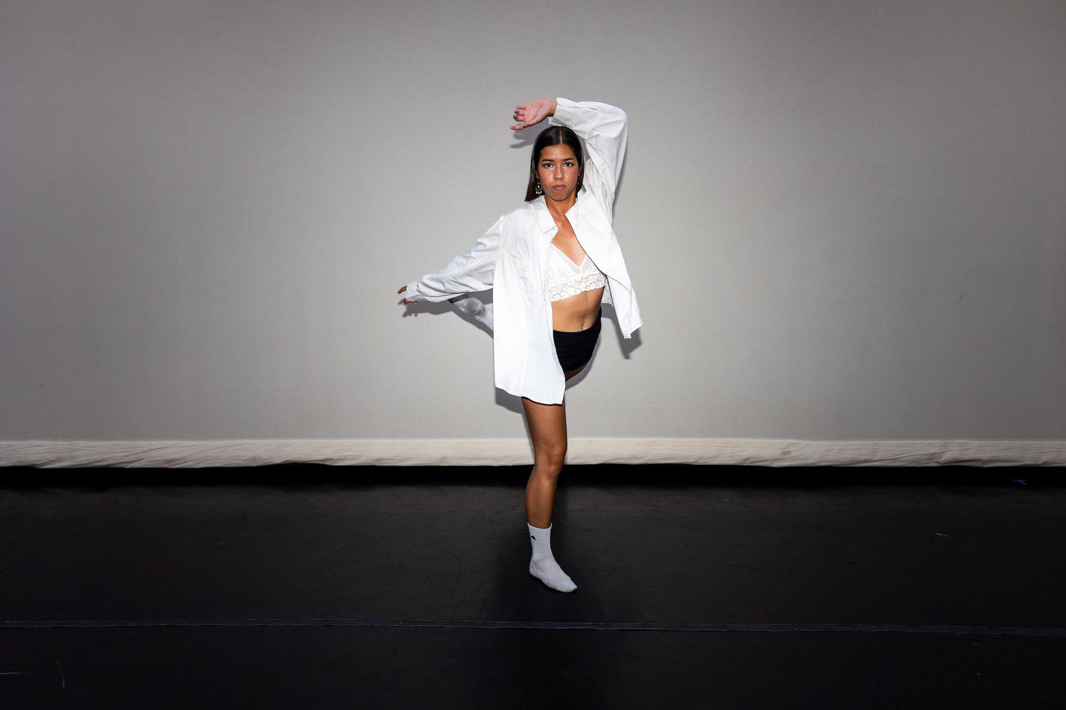 A dancer in a white shirt and black shorts performs a high kick on a dark studio floor.
