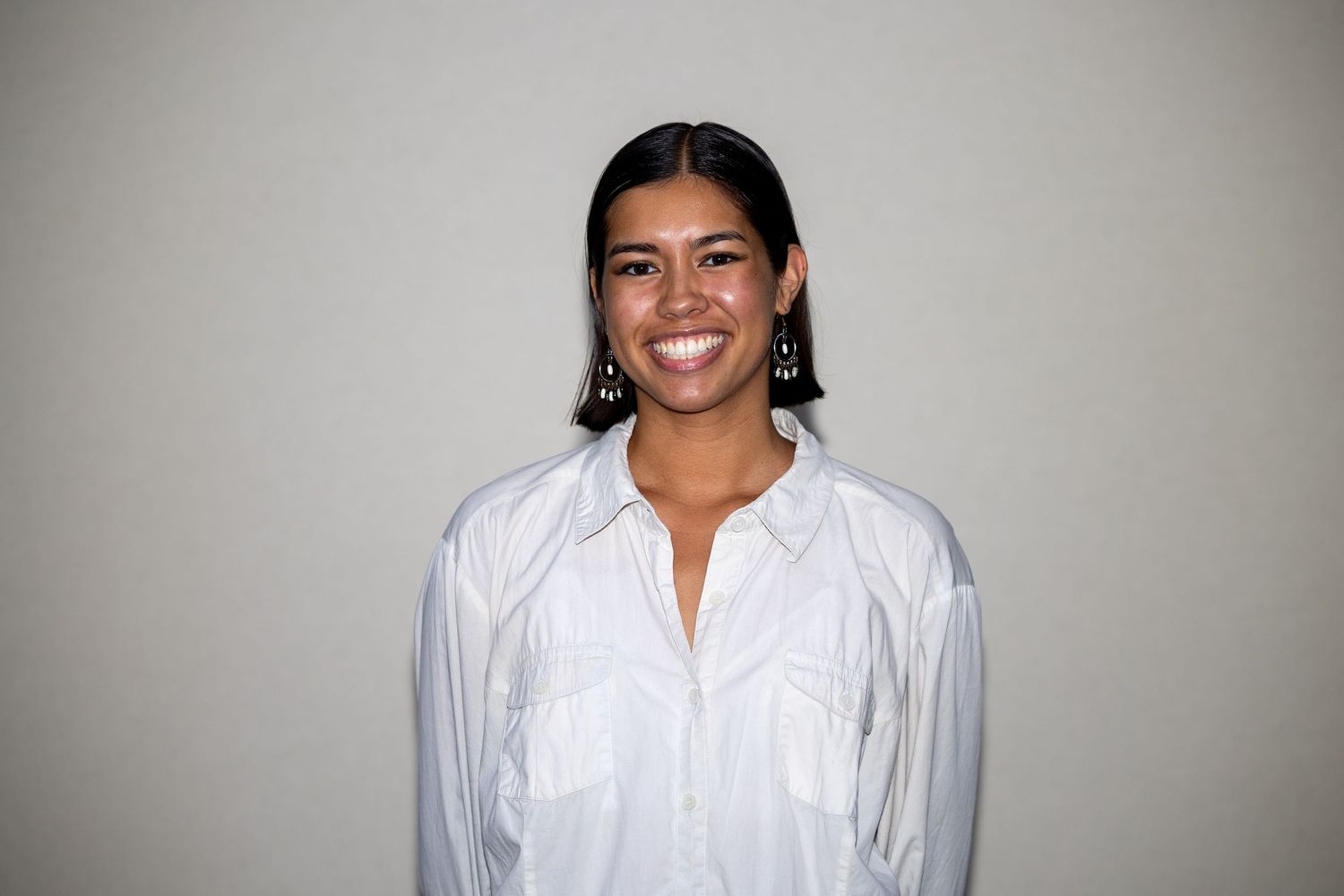 Happy woman wearing white oversized shirt and black earrings smiling confidently against gray backdrop.