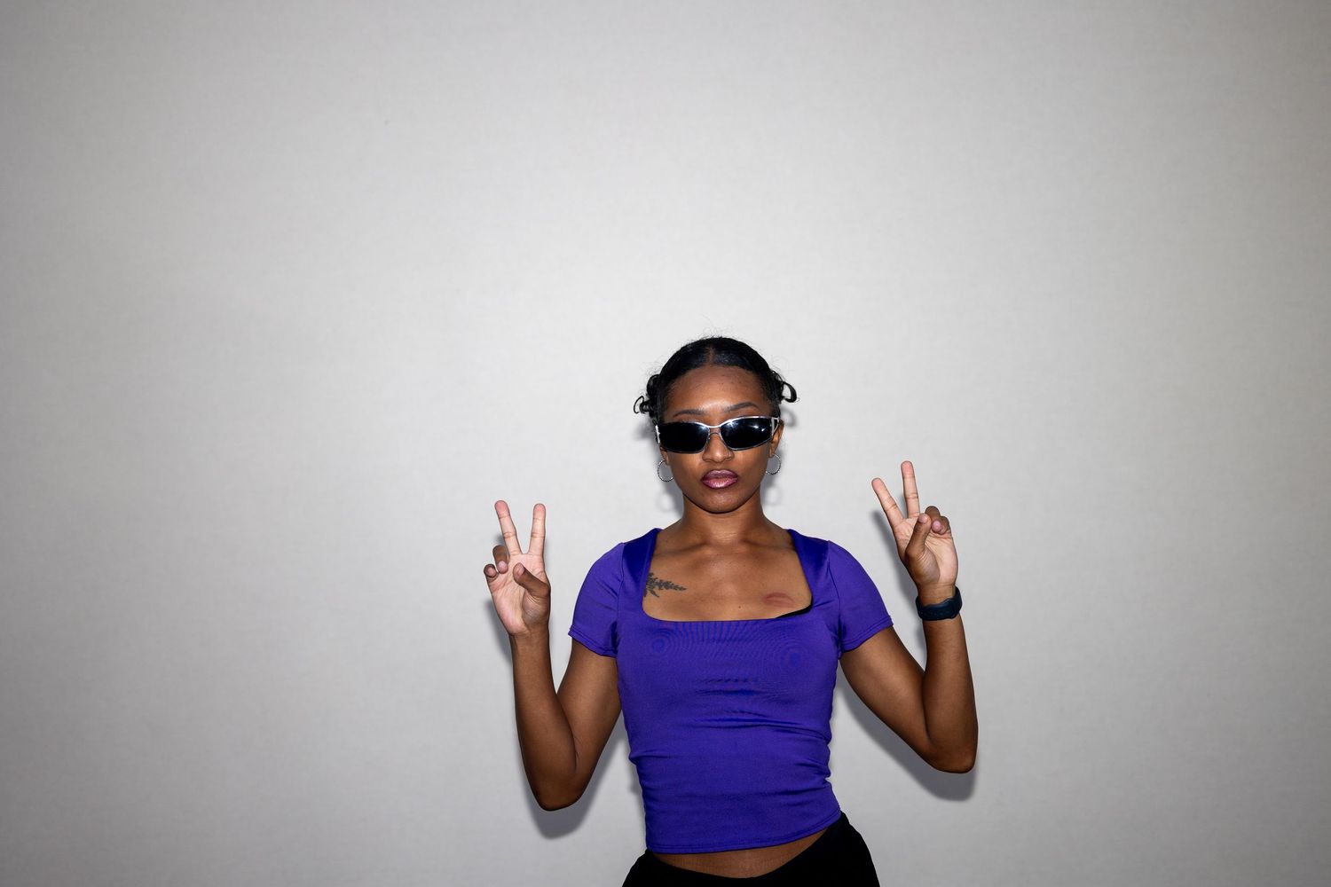 A stylish woman in a purple crop top and sunglasses flashes peace signs against a white background.