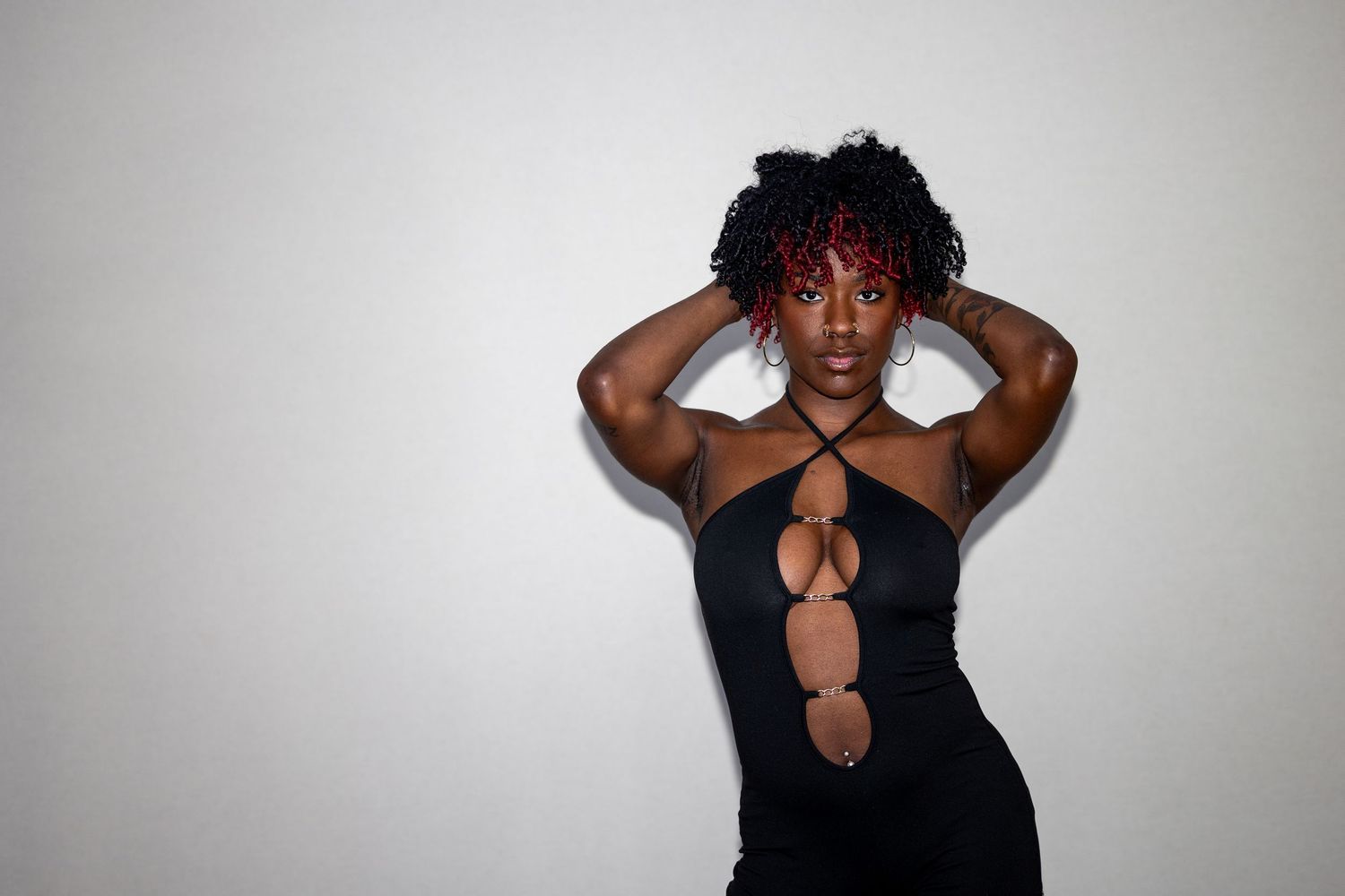 Woman in black cutout dress posing with hands behind head against white background.