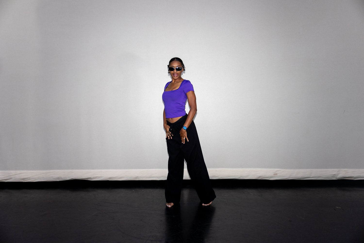 Stylish woman in purple tee and wide-leg black pants wearing sunglasses on stage.