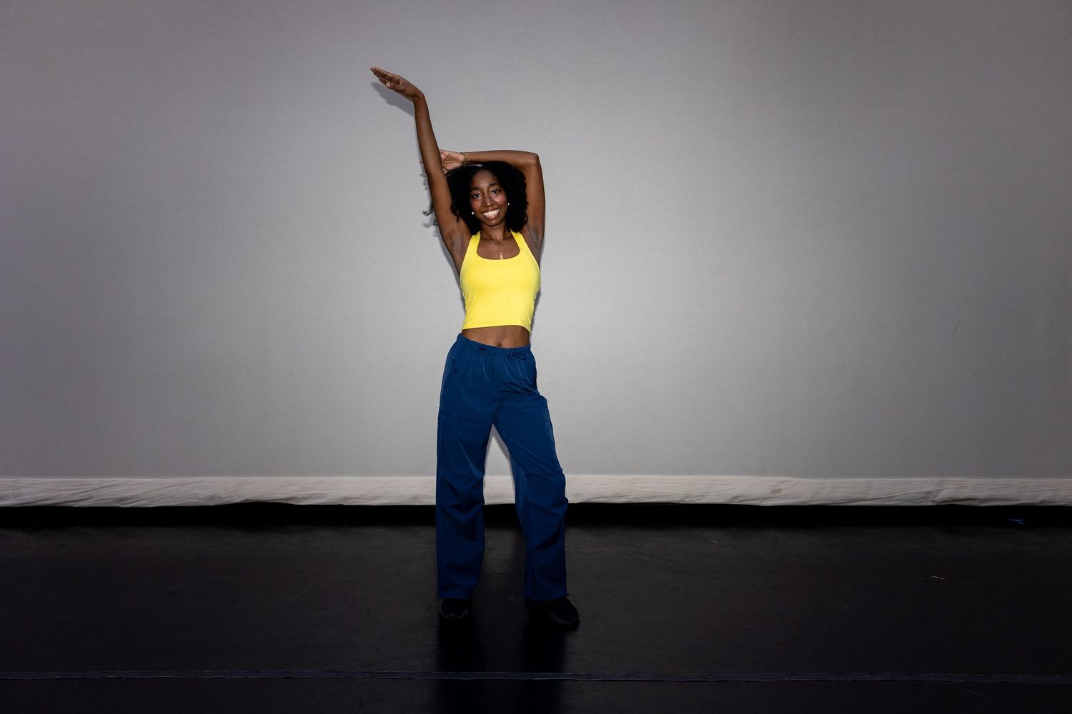 A woman in a yellow crop top and blue wide-leg pants dancing gracefully against a gray background.