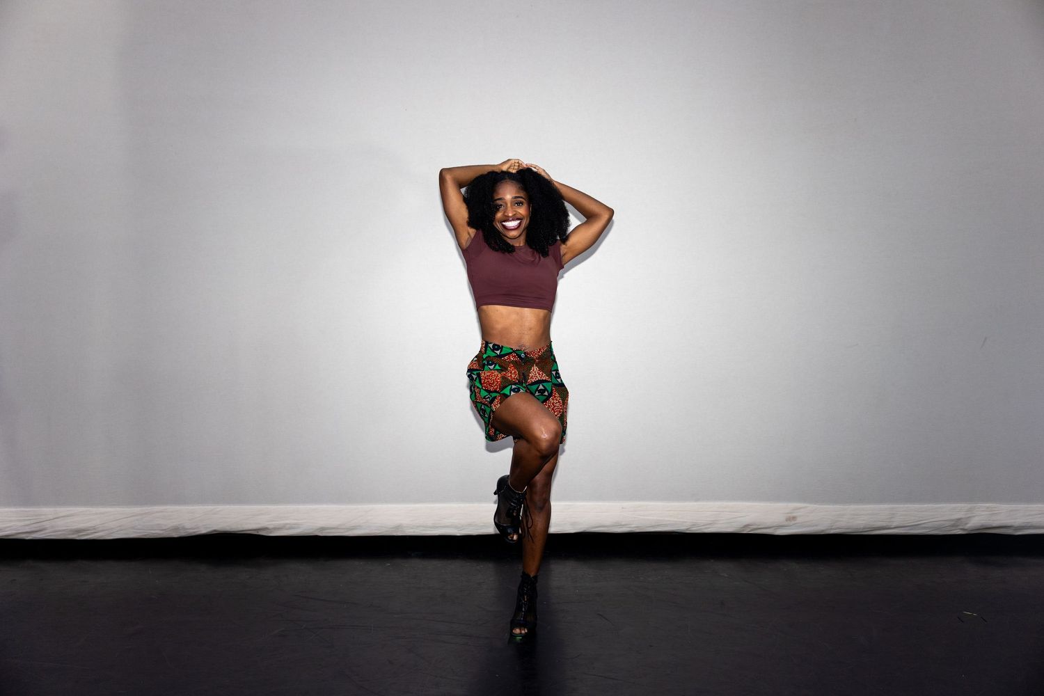Black female dancer smiles while posing in maroon crop top and colorful African print shorts.