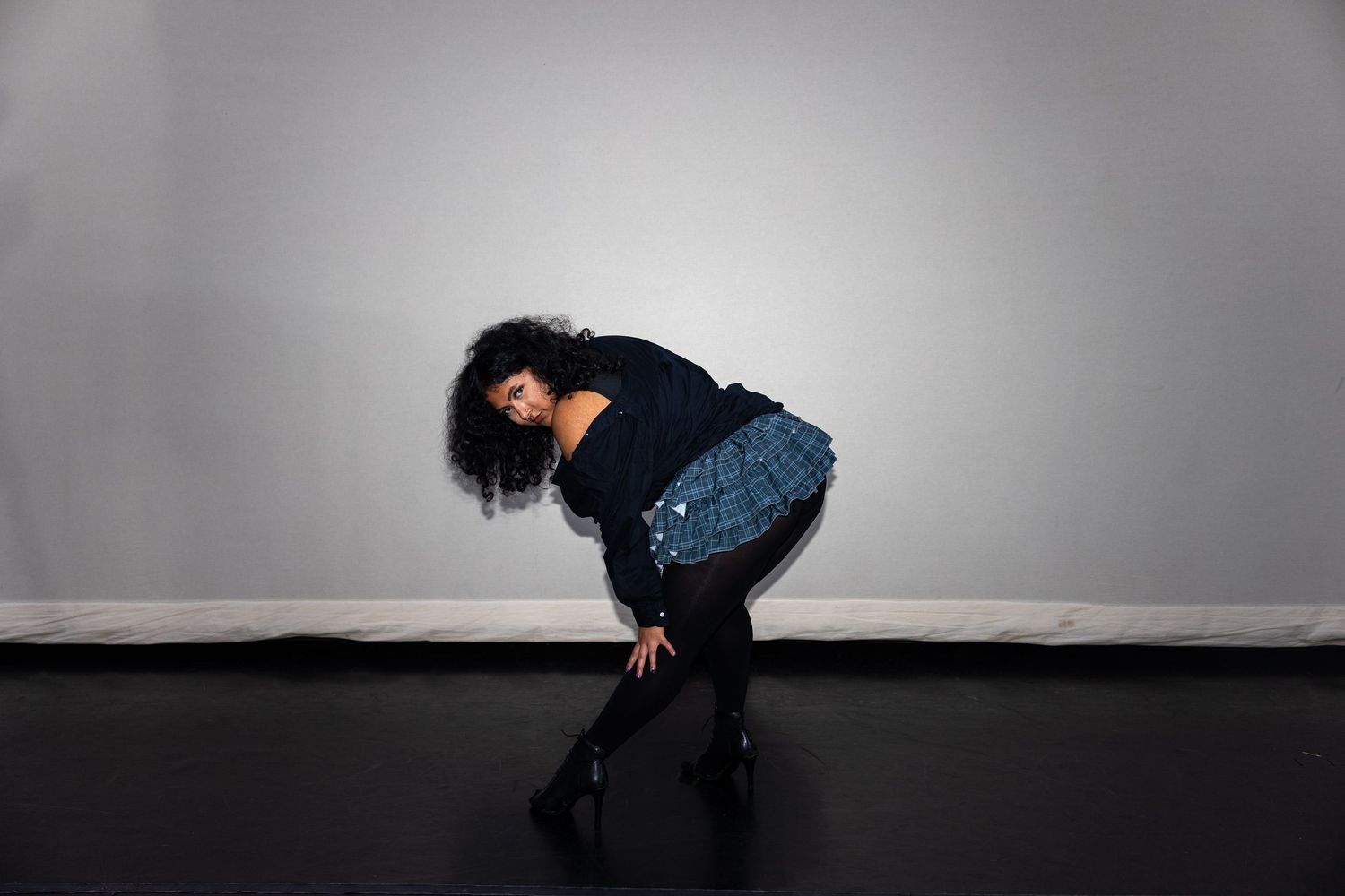 A dancer bends sideways in a blue plaid skirt and black top on a dark studio floor.