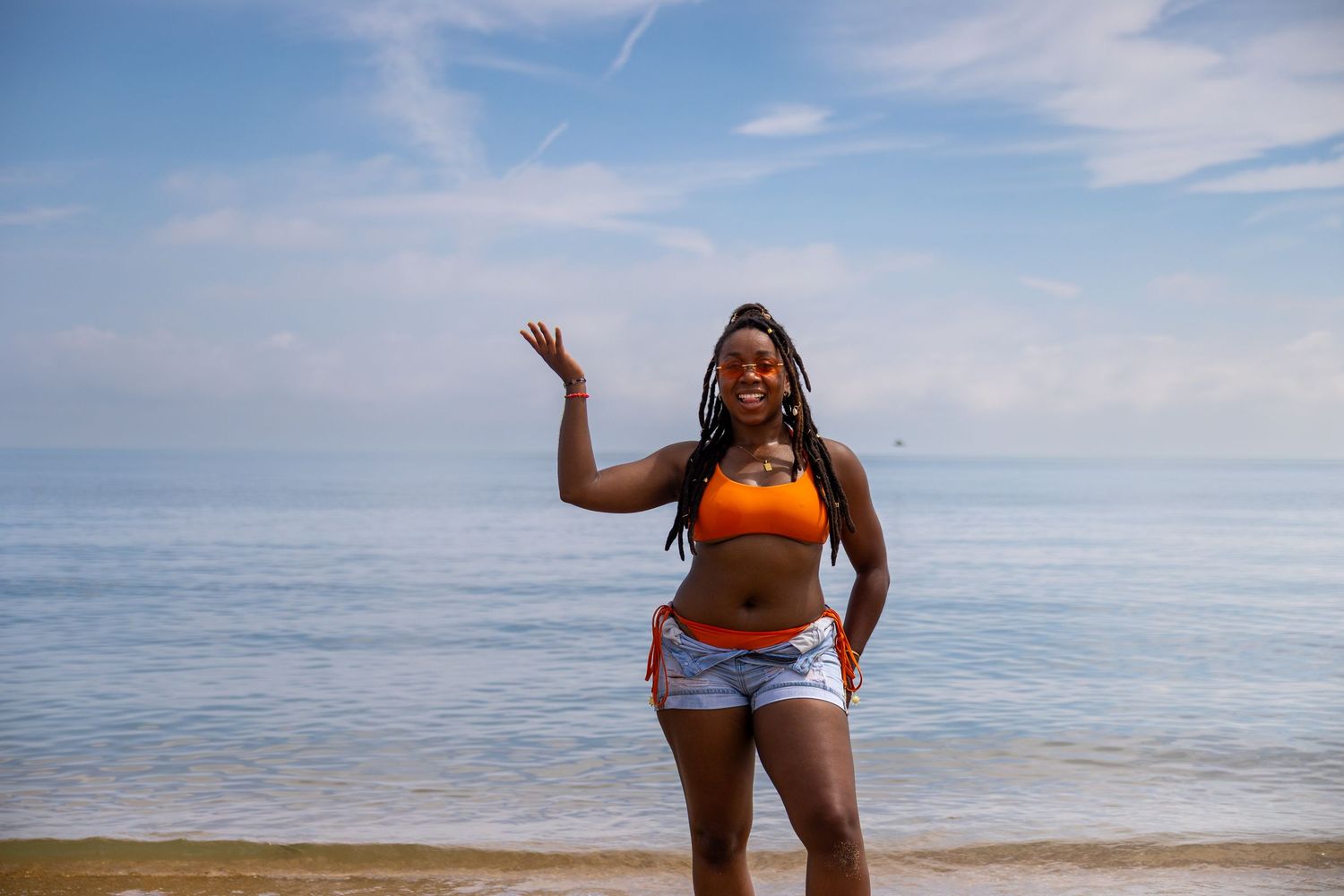 A smiling woman in an orange bikini top and denim shorts poses on a sunny beach with blue ocean behind her.