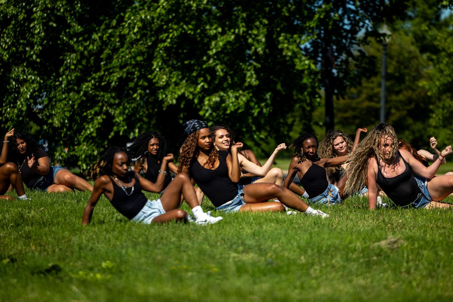 Group of women in black outfits sitting on green grass in a sunny park surrounded by lush trees.