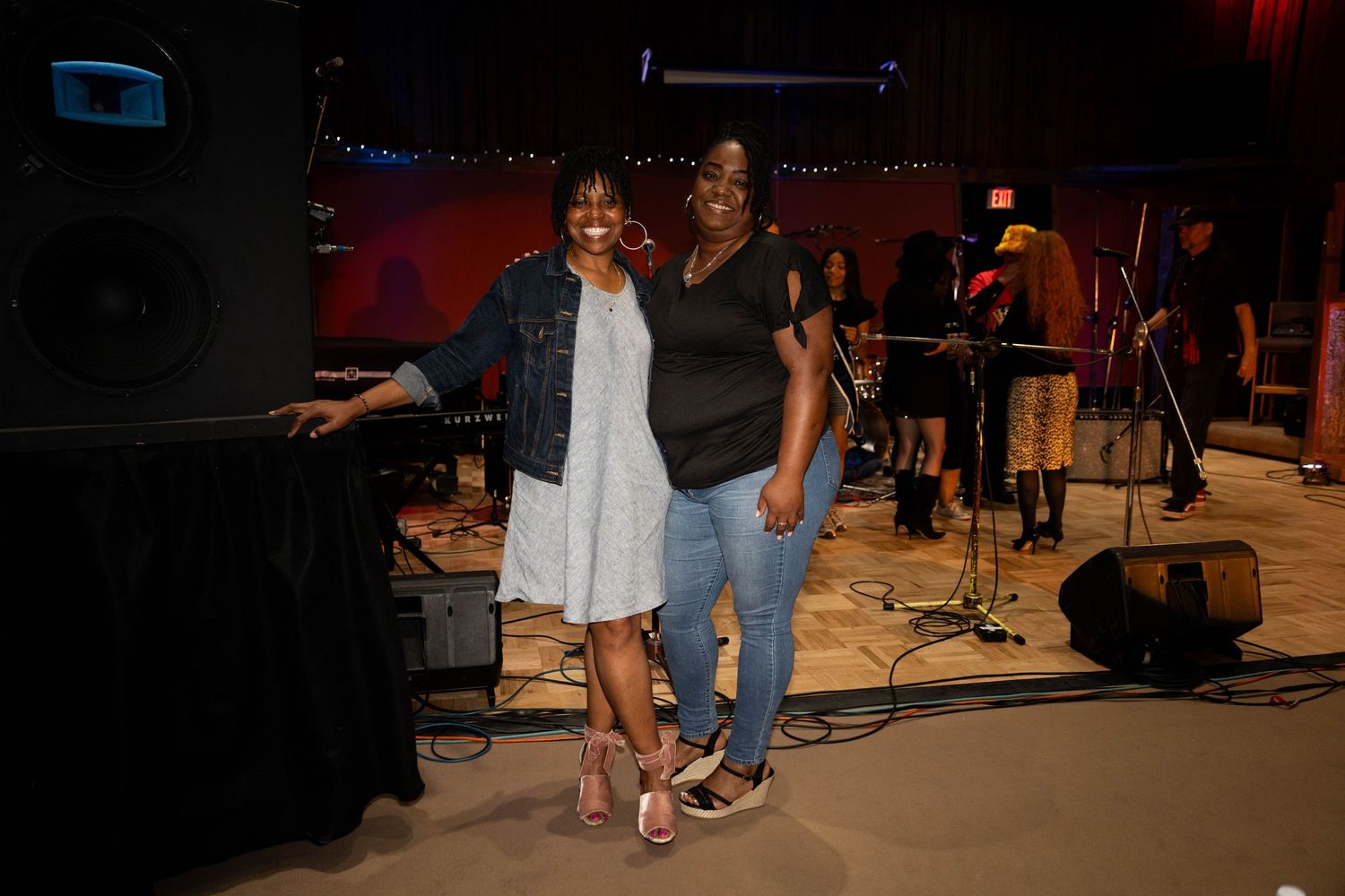 Two women smile and pose together near a speaker at an indoor music venue event.