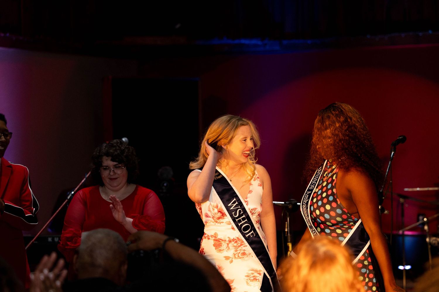 Two women wearing pageant sashes on stage at an indoor event with band members visible in background.