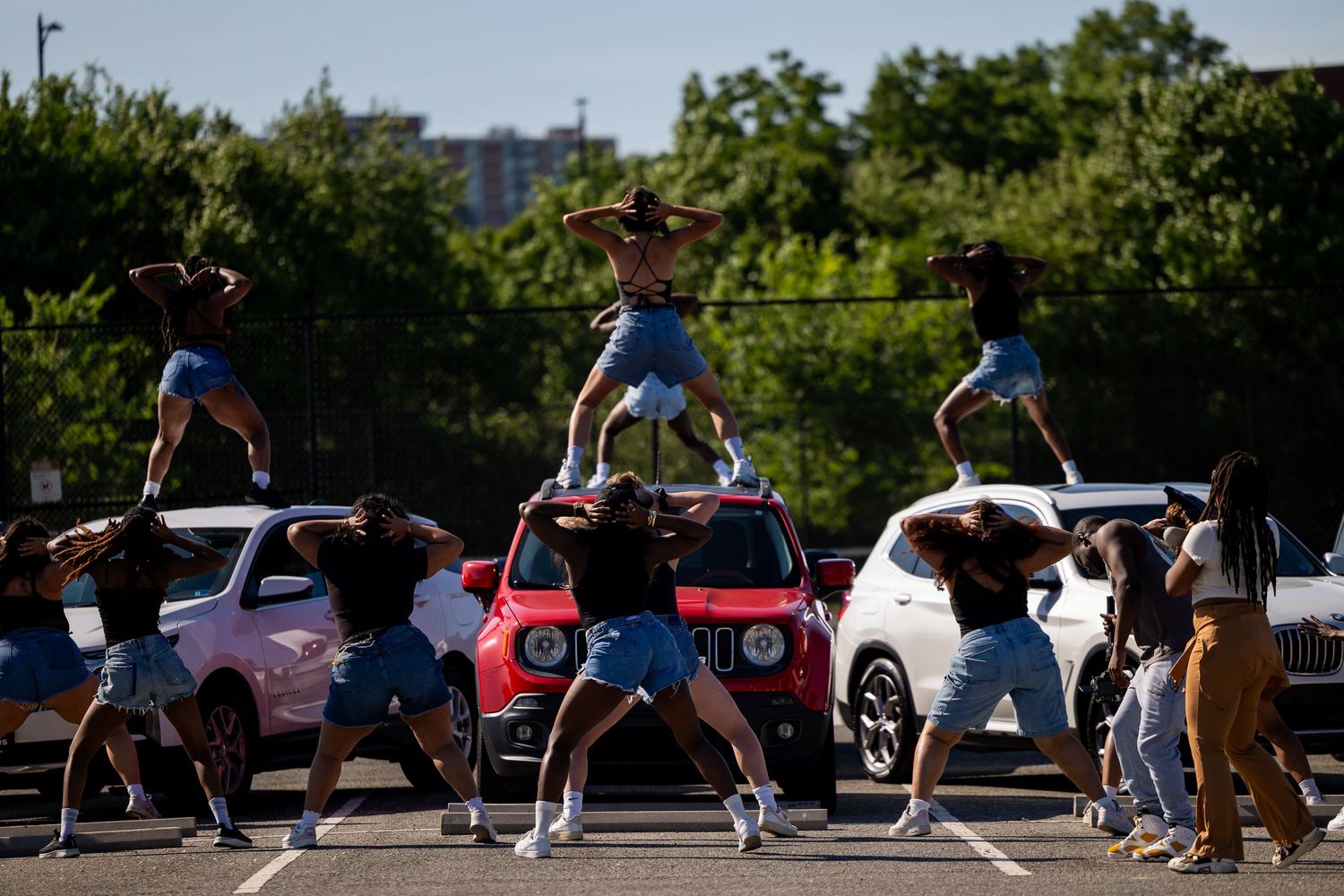 A cheerleading squad forms a human pyramid in a parking lot, wearing denim shorts and black tops.