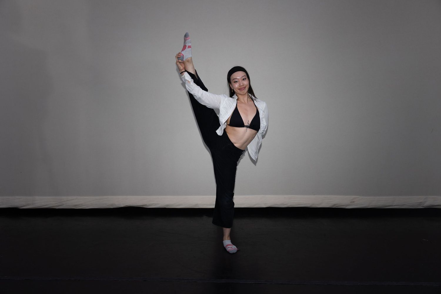Flexible dancer in black pants and white jacket performs high kick while holding a ribbon in a studio.