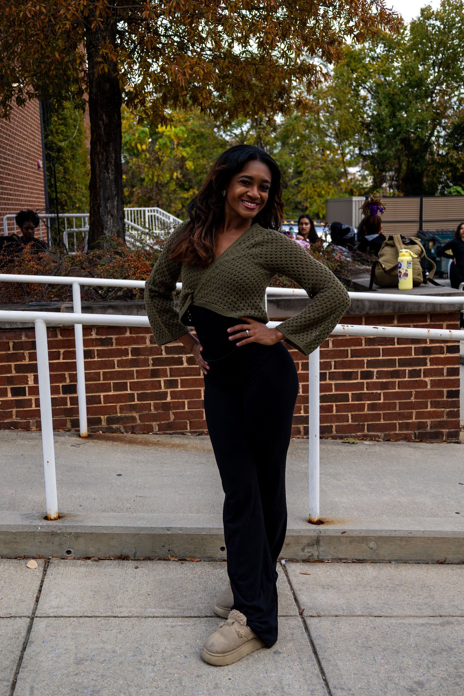 Person in dark clothing strikes a playful pose against a brick wall with white railing during autumn.