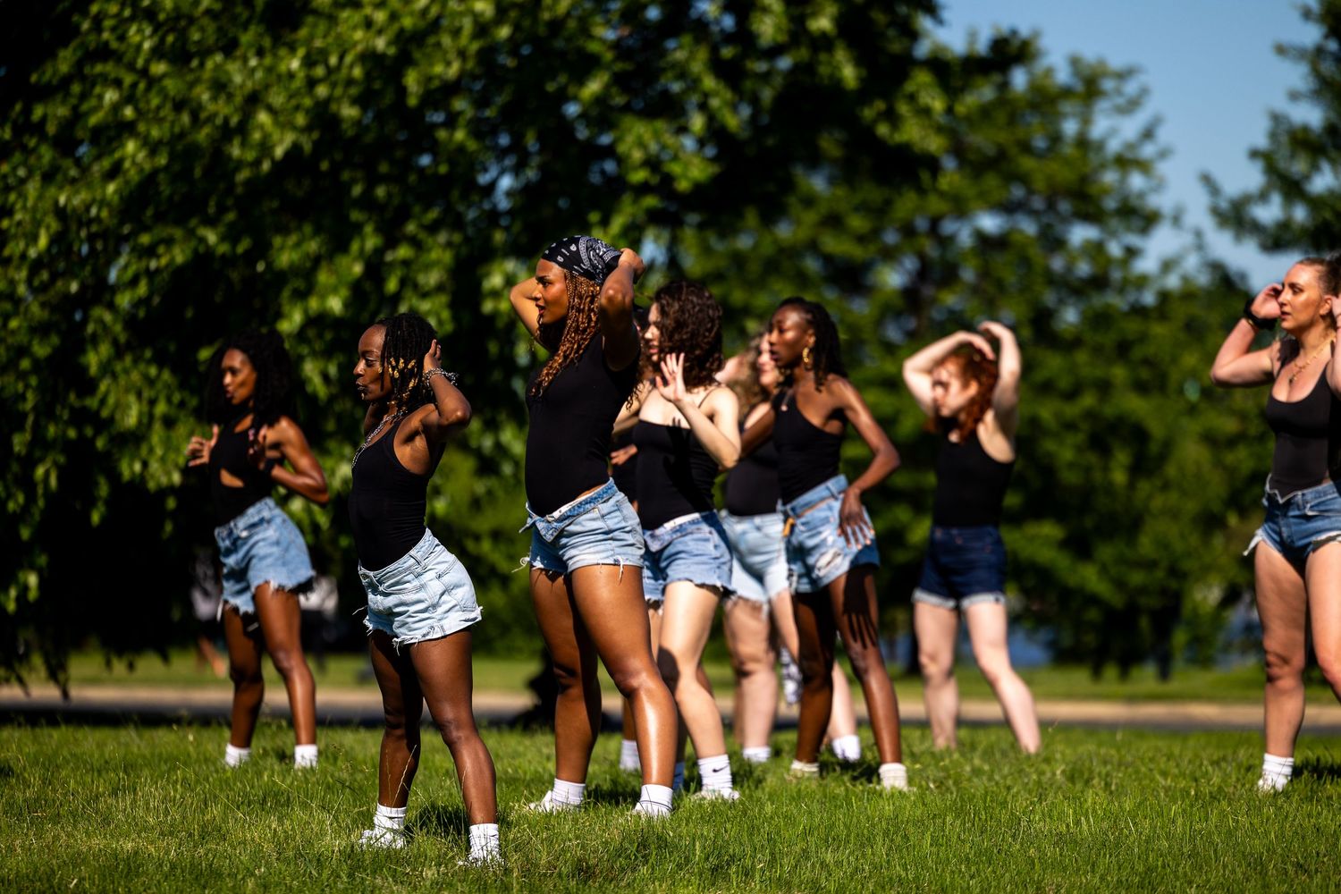 A group of young women in black tops and denim shorts performing a synchronized dance routine outdoors in a park.