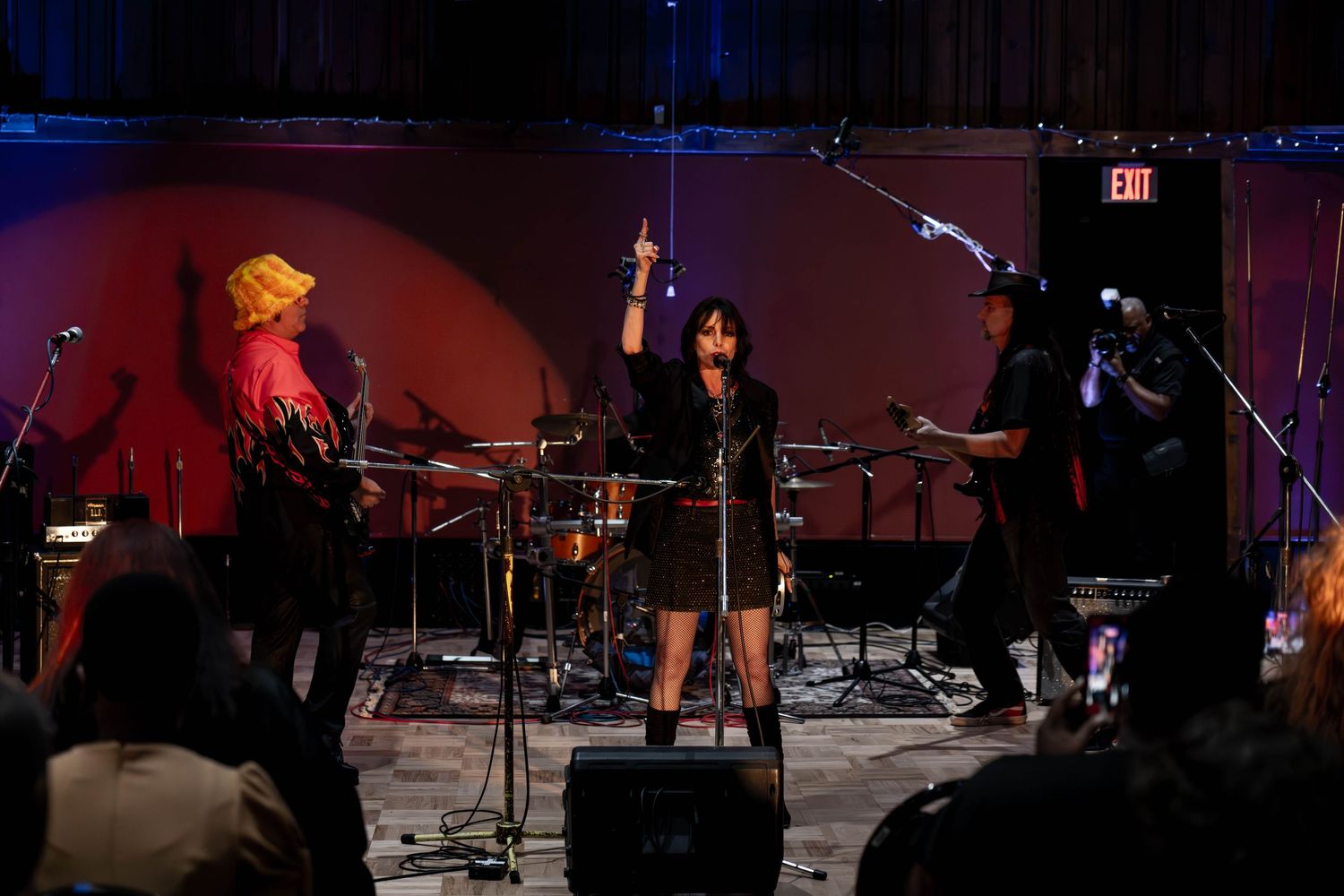 Live band performance on stage with singer raising hand, drummer, and guitarist under red and blue lights.