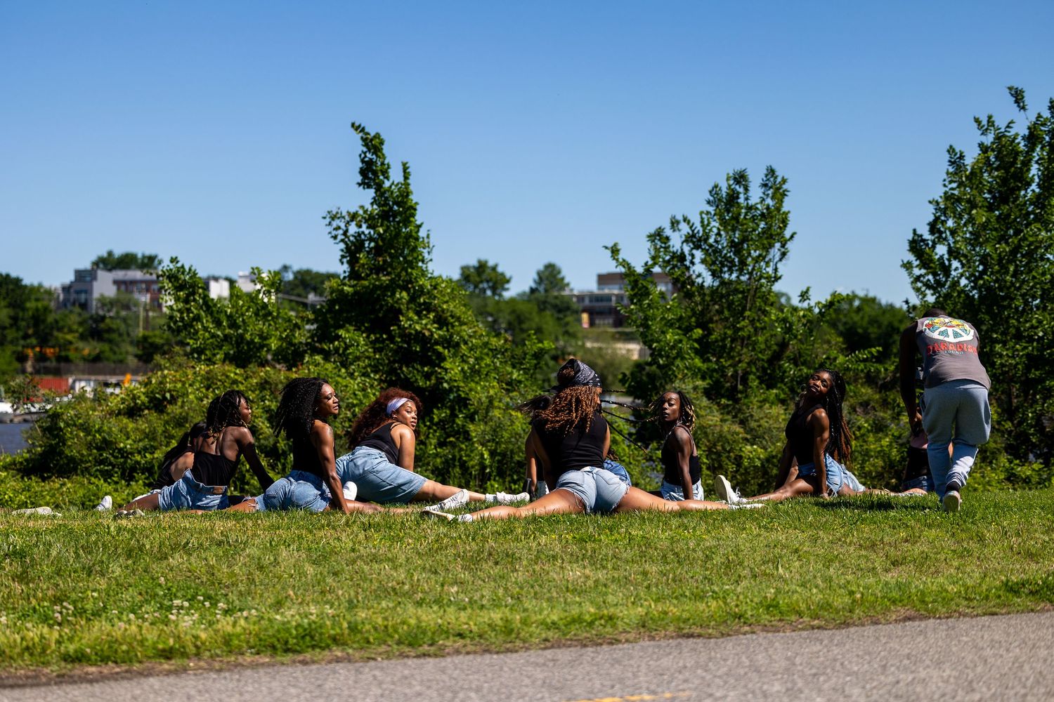 Group of young women sitting on green grass in a sunny park with trees and blue sky in the background.