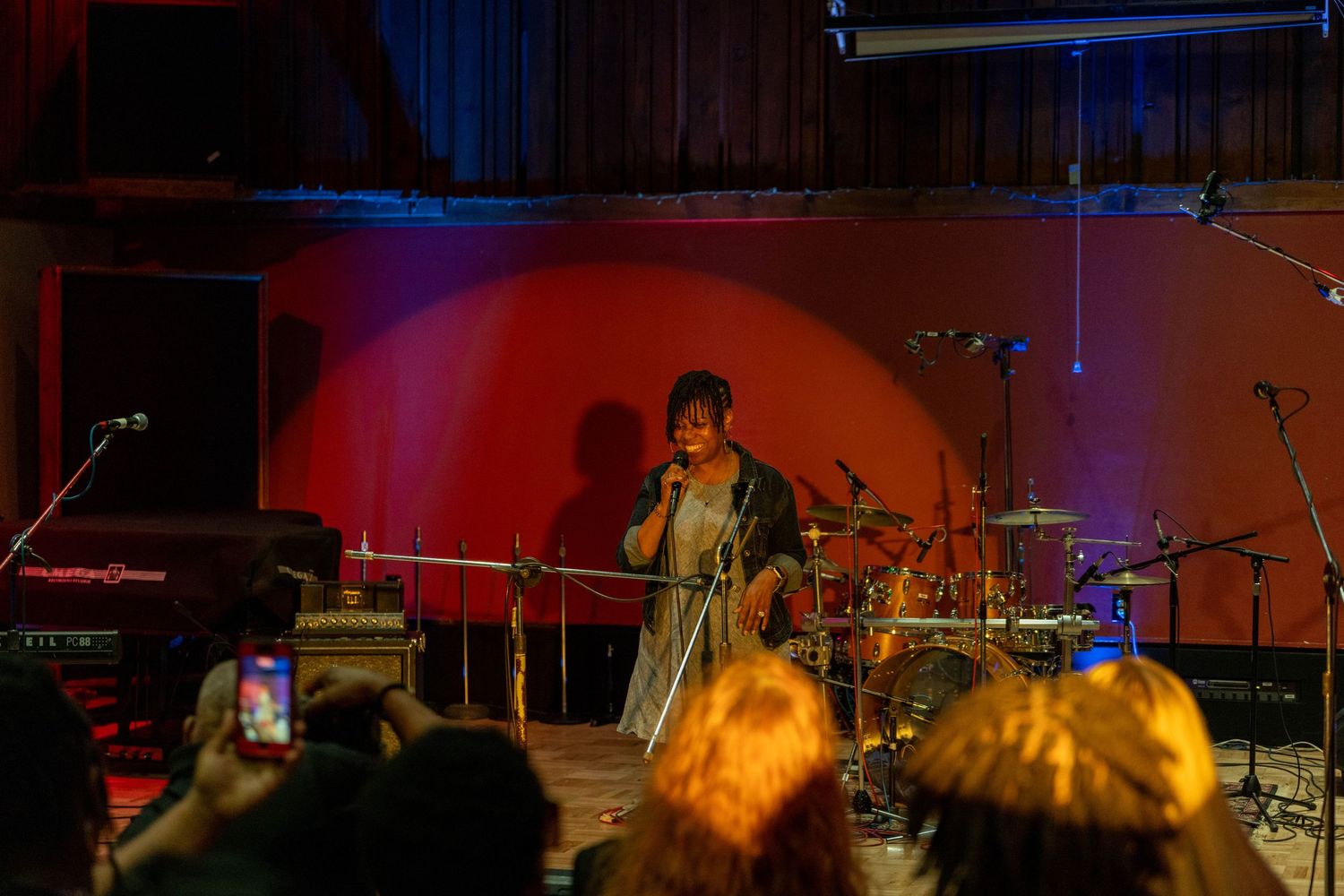 Singer performing on stage before a crowd, red spotlight background, audience filming with phones.
