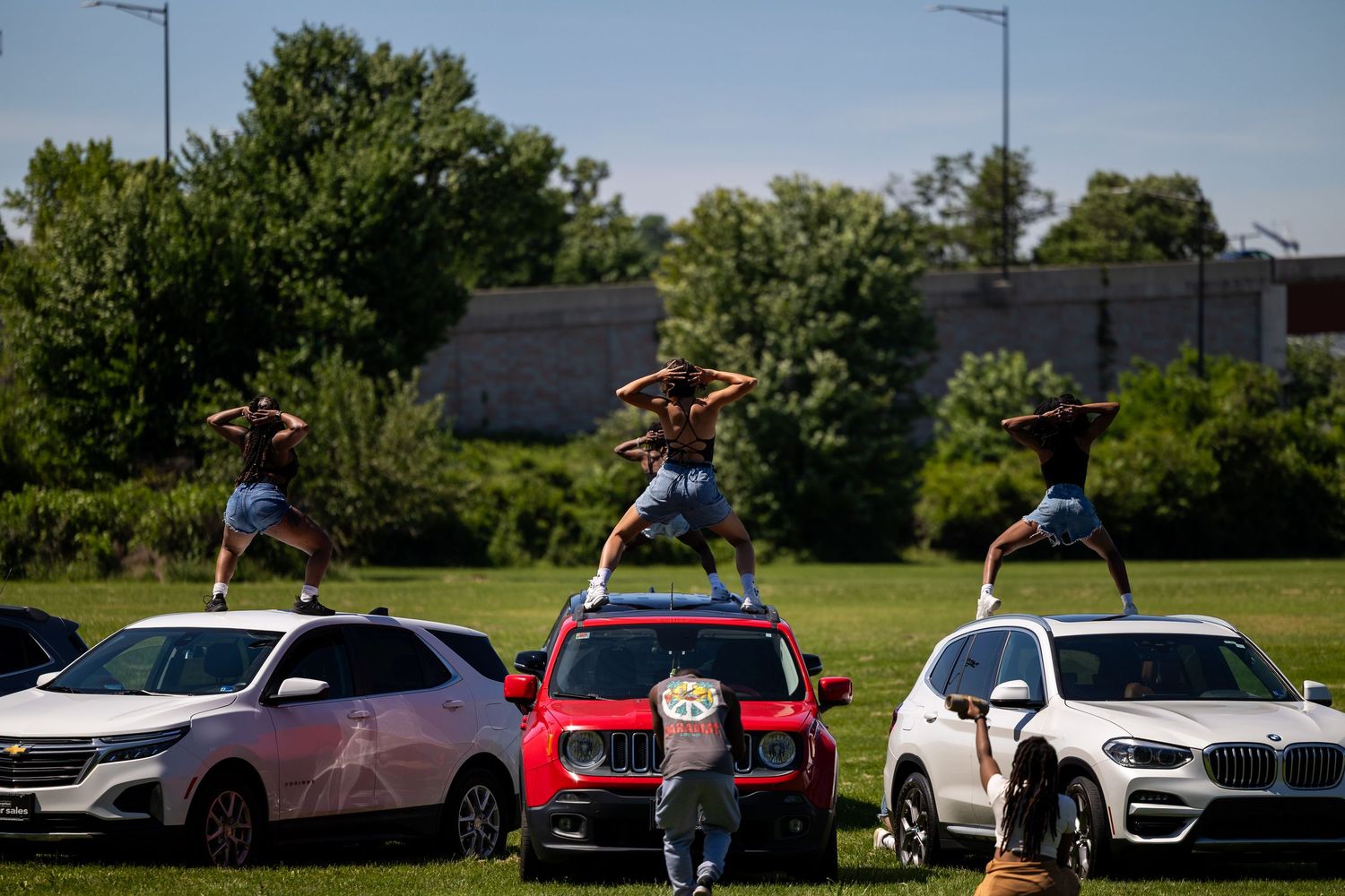 Dancers perform atop cars outdoors at a drive-in event, with green trees and a brick wall in the background.