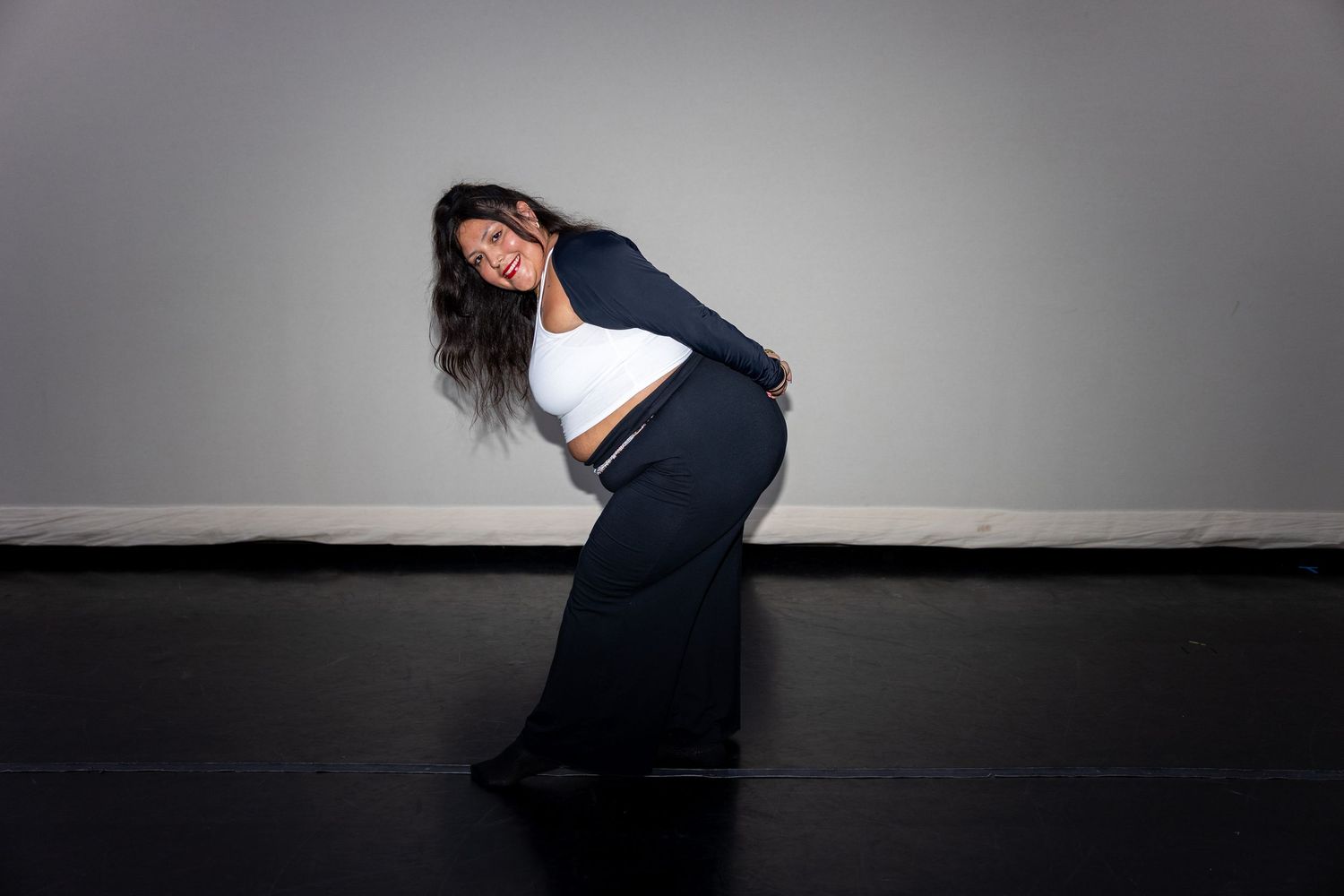 Woman in white crop top and navy wide-leg pants poses gracefully in a dance studio, smiling with flowing hair.