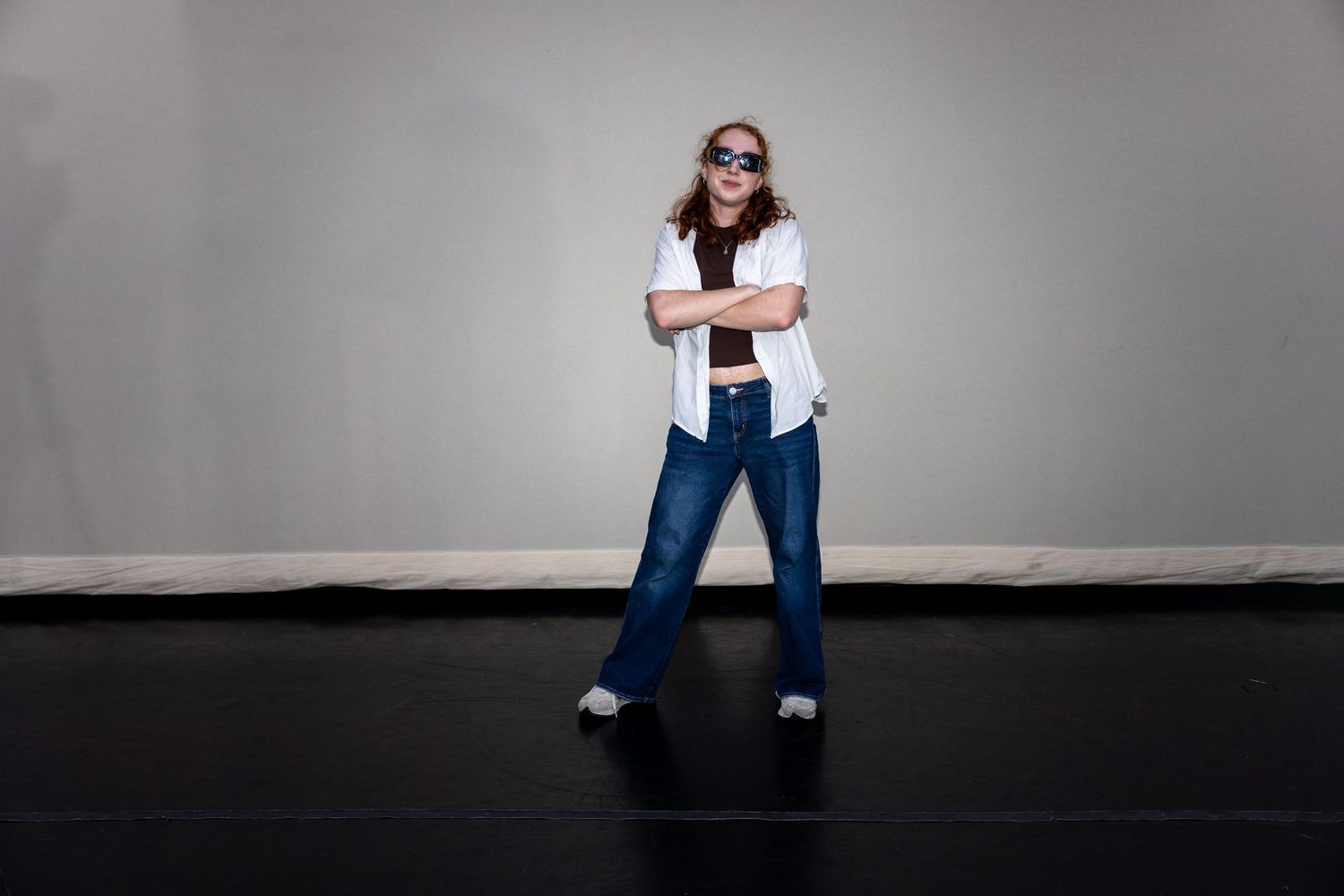 A young woman in jeans, white shirt, and sunglasses stands with arms crossed on a dark dance studio floor.