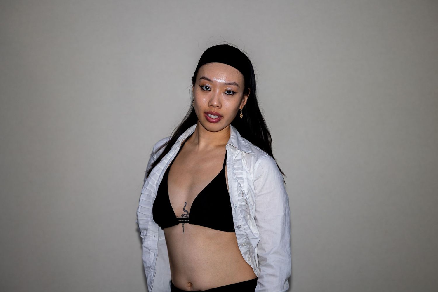 Young Asian woman wearing a black bralette and open white jacket, posing with a confident expression.