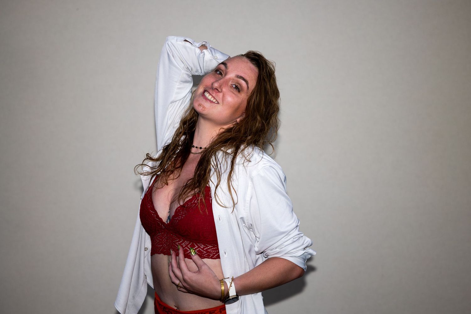 A smiling woman with wavy hair poses playfully in a red lace bralette and open white shirt.
