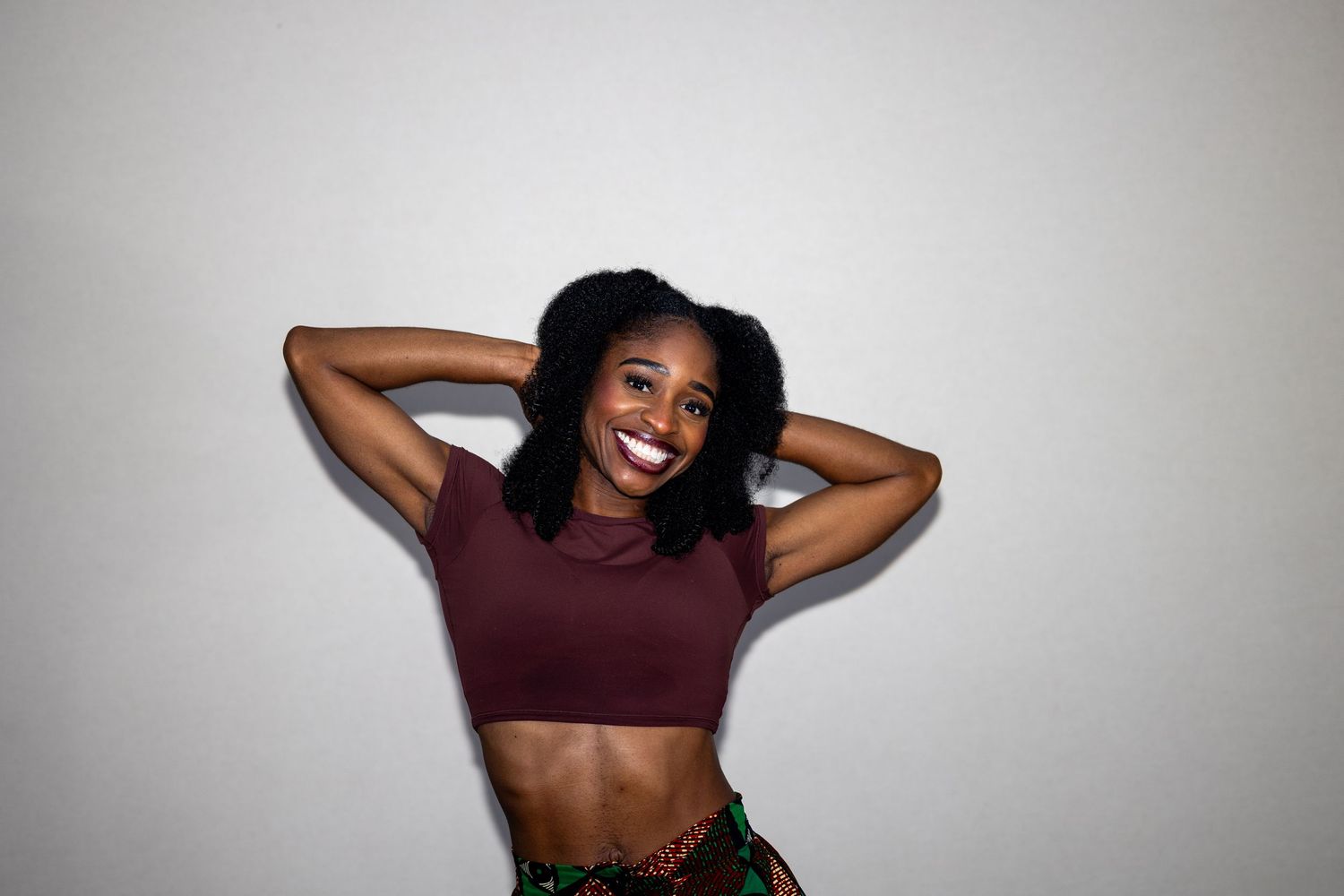 Happy Black woman in maroon crop top and printed skirt posing with hands behind her head.