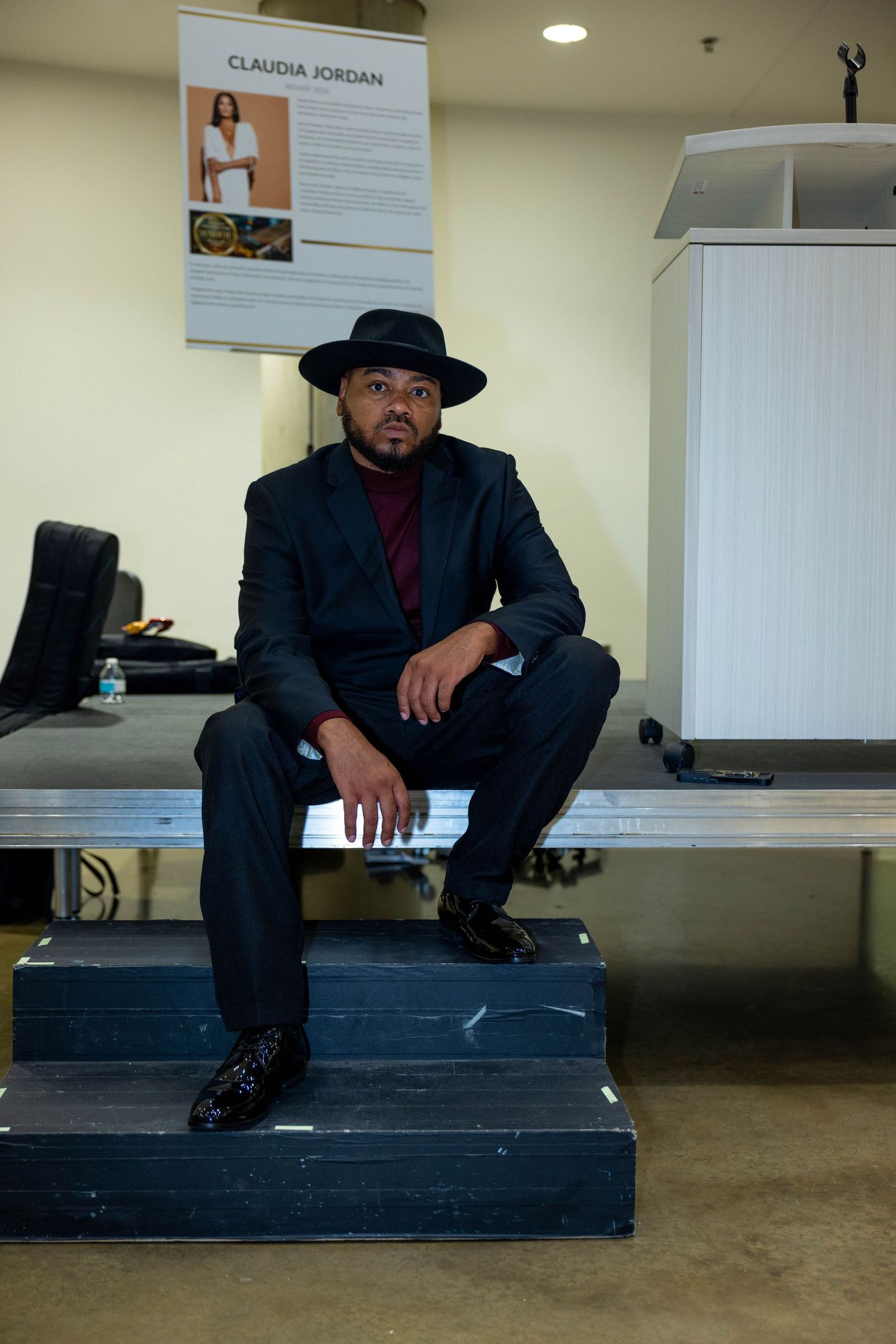 A well-dressed man in a dark suit and black hat sits confidently on steps inside an office space.