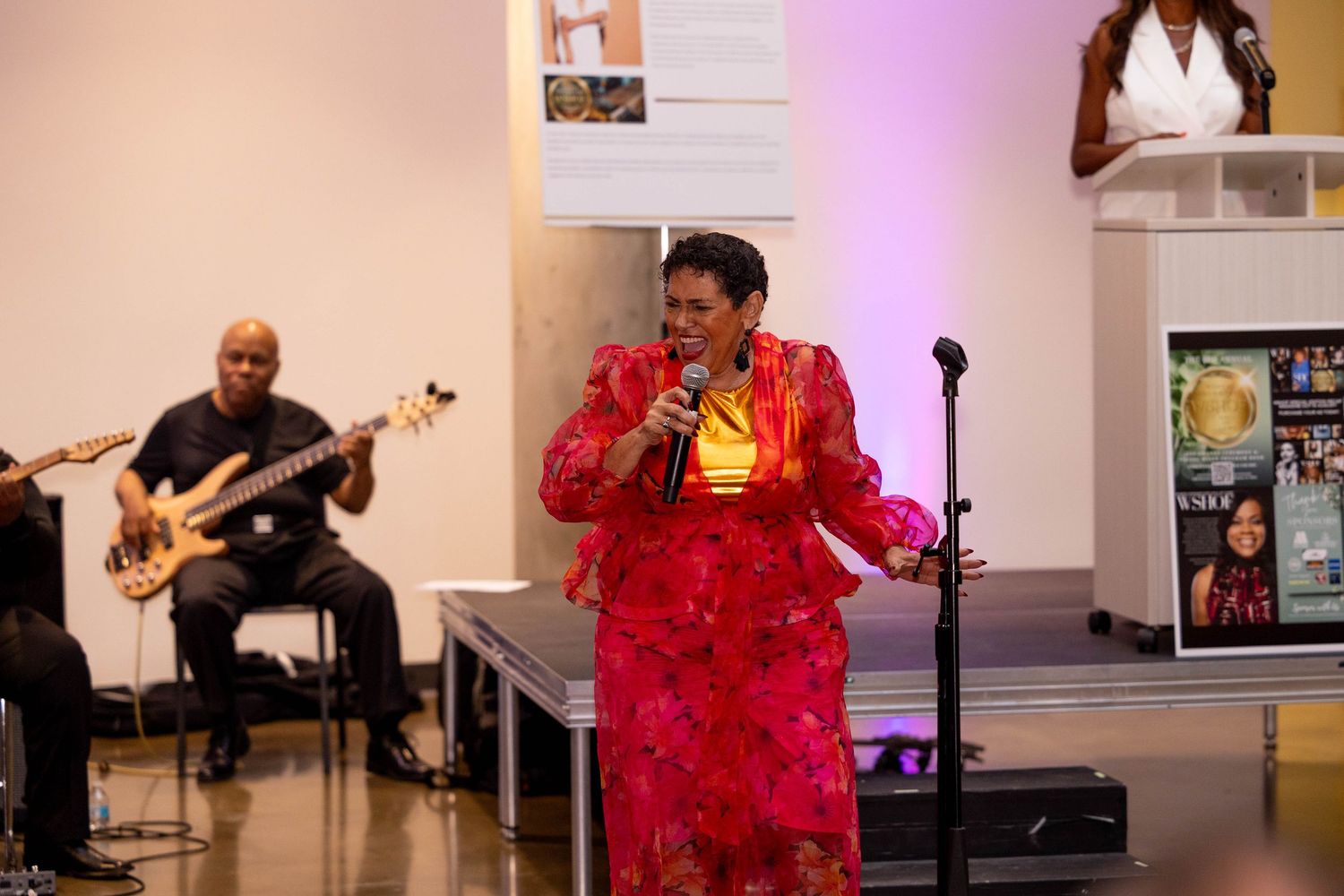 Woman in red floral dress performing on stage with microphone while bassist plays guitar in background.